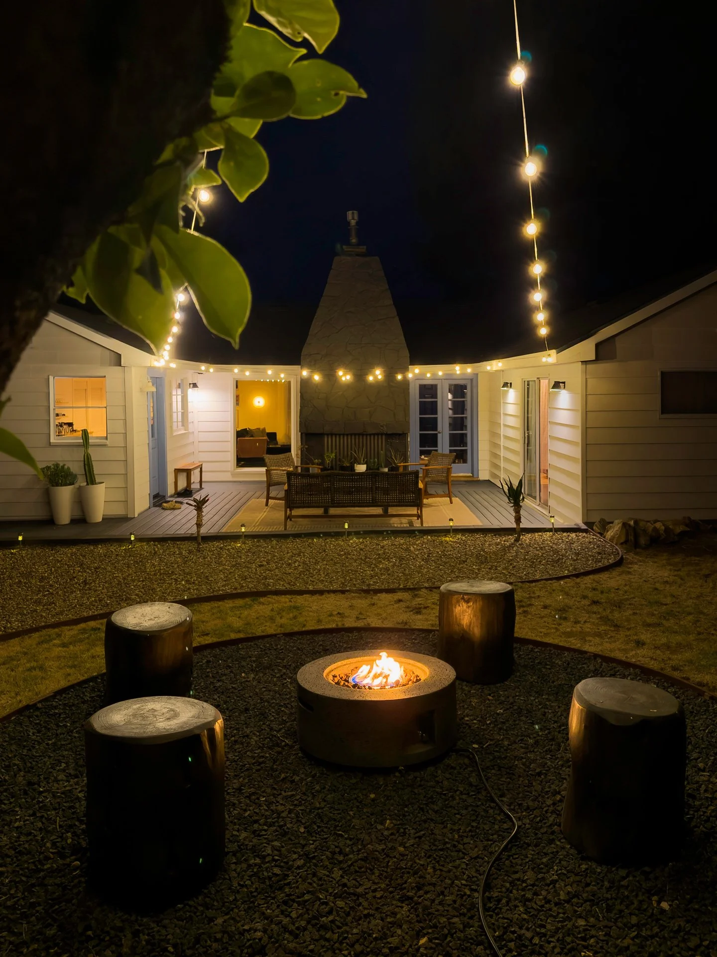 Nighttime outdoor patio scene with a fire pit surrounded by tree stump seats, string lights hanging above, a house with a fireplace, and a deck with outdoor furniture.