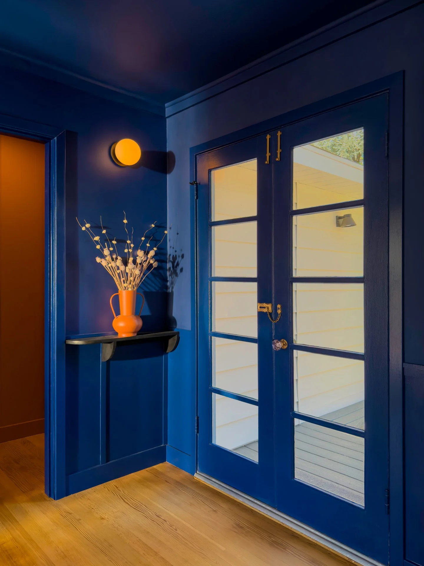 A blue front door with glass panels opens to an outdoor porch, with a yellow wall and a wall lamp visible outside. Inside, there's a blue wall with a small shelf holding an orange vase with decorative white branches, a blue wall-mounted light, and a wooden floor.