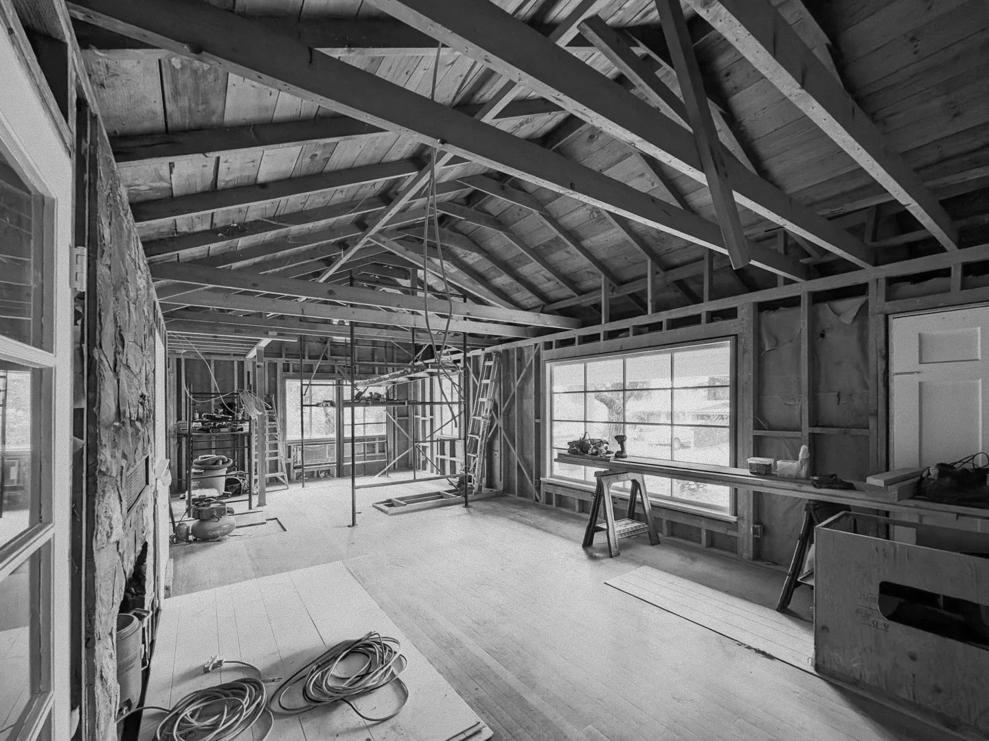 Interior view of a room under construction, showing exposed wooden walls, ceiling beams, construction tools, supplies, and a ladder, with large windows and a door.