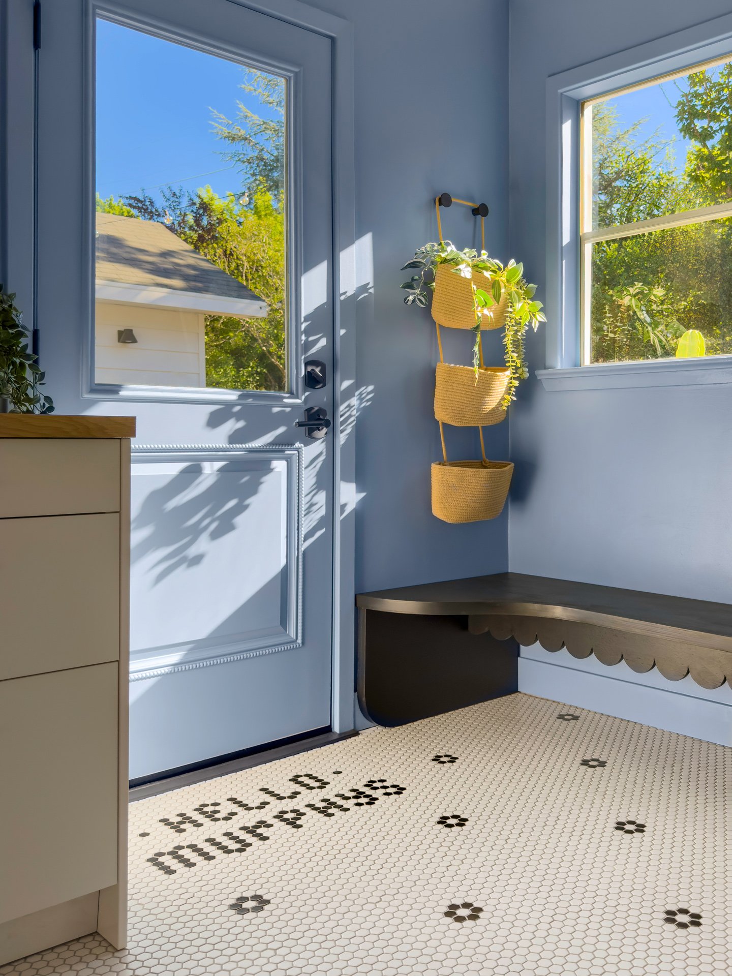 Interior view of a sunlit porch with a white door, large windows showing trees and a house roof outside, a beige cabinet, and a black and white tiled floor with a decorative pattern spelling 'BONUS'.