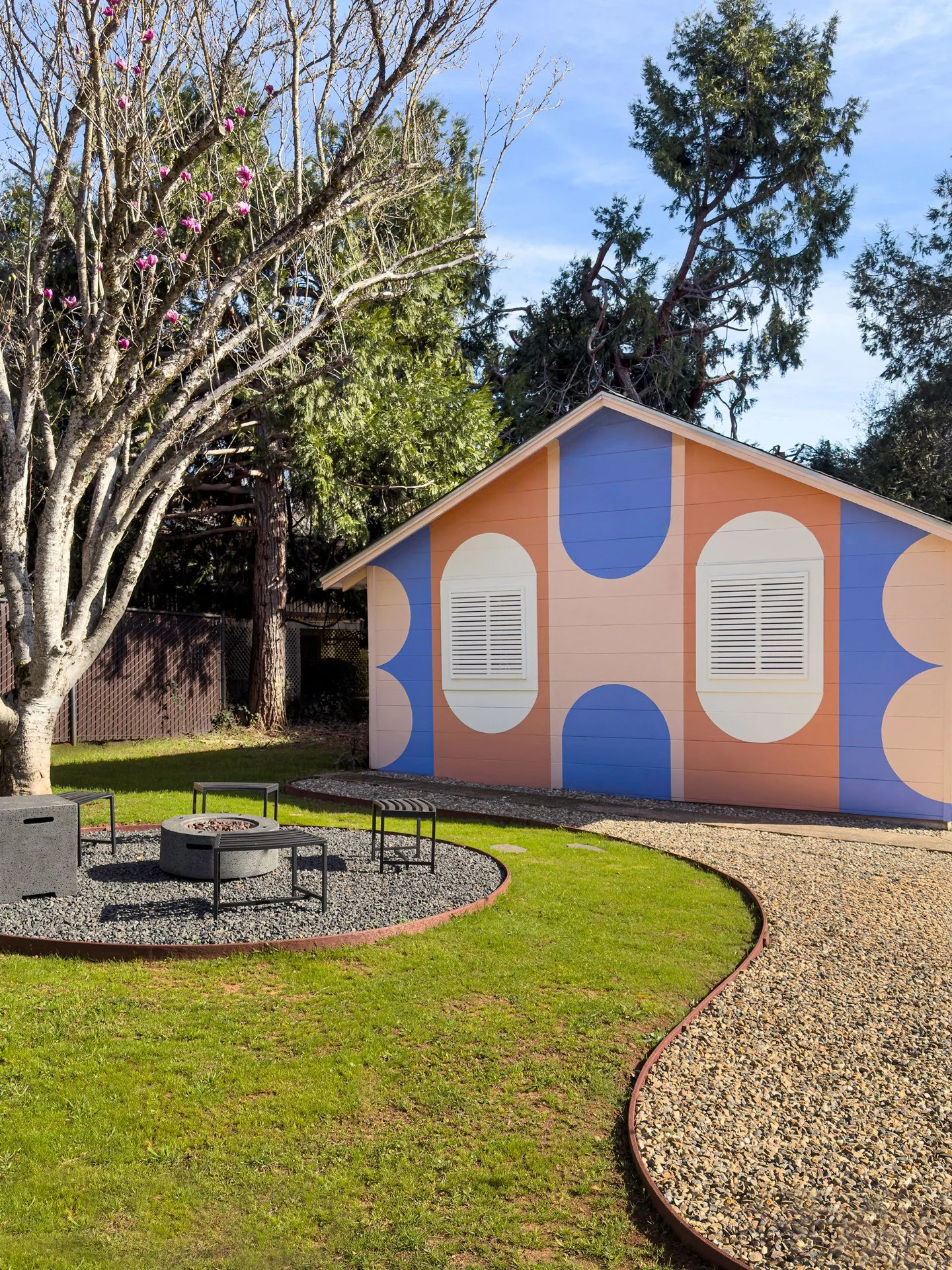 A colorful small building with blue and orange geometric patterns on the exterior wall, a sloped roof, set in a park with a fire pit, chairs, and a curved gravel pathway surrounded by green grass and trees.