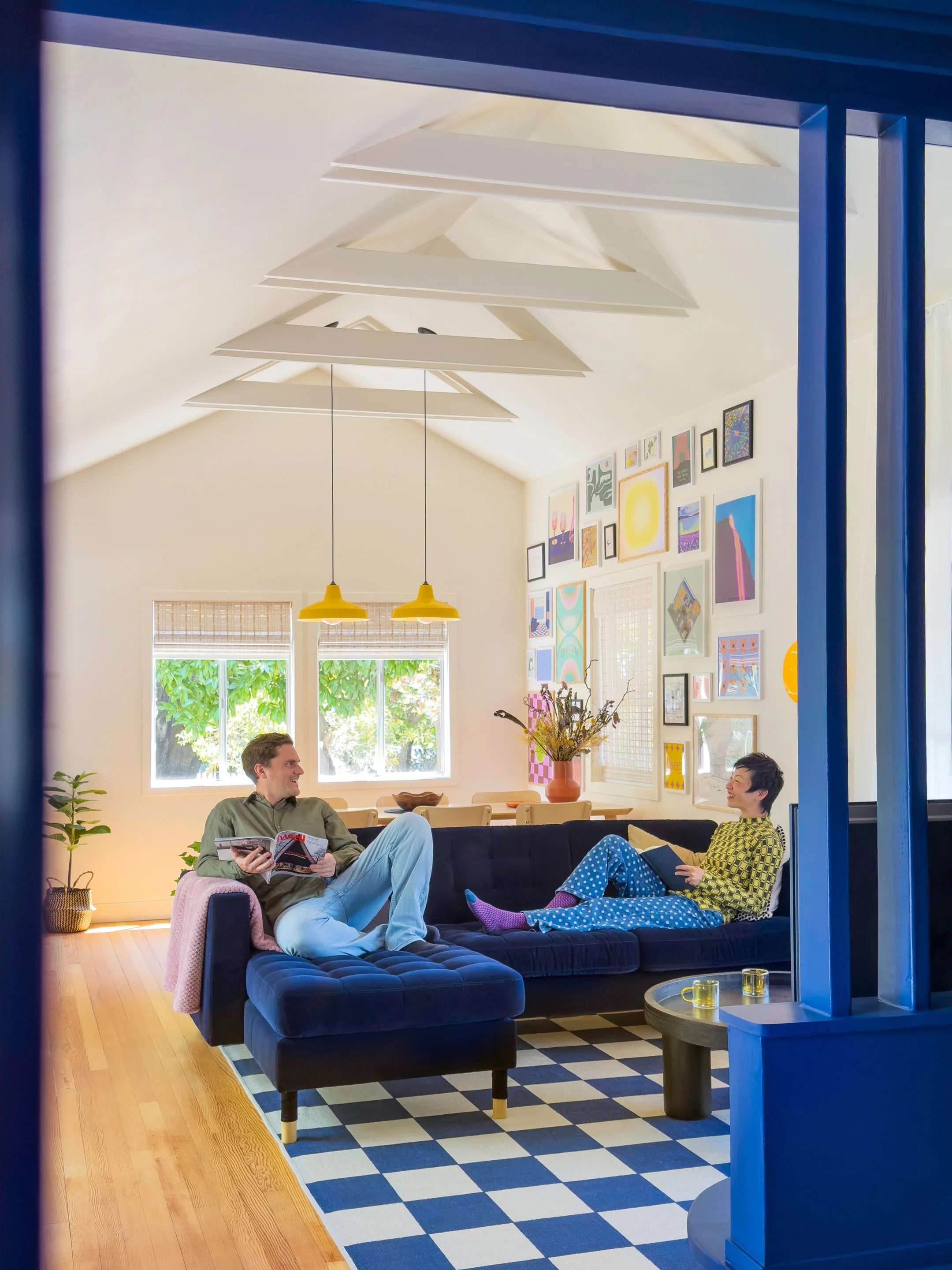 Two people sitting on a dark blue sofa in a bright living room, chatting and smiling. The man is holding a magazine, and the woman is holding a tablet. The room has modern decor with colorful wall art, yellow pendant lights, large windows, and a checkered rug.