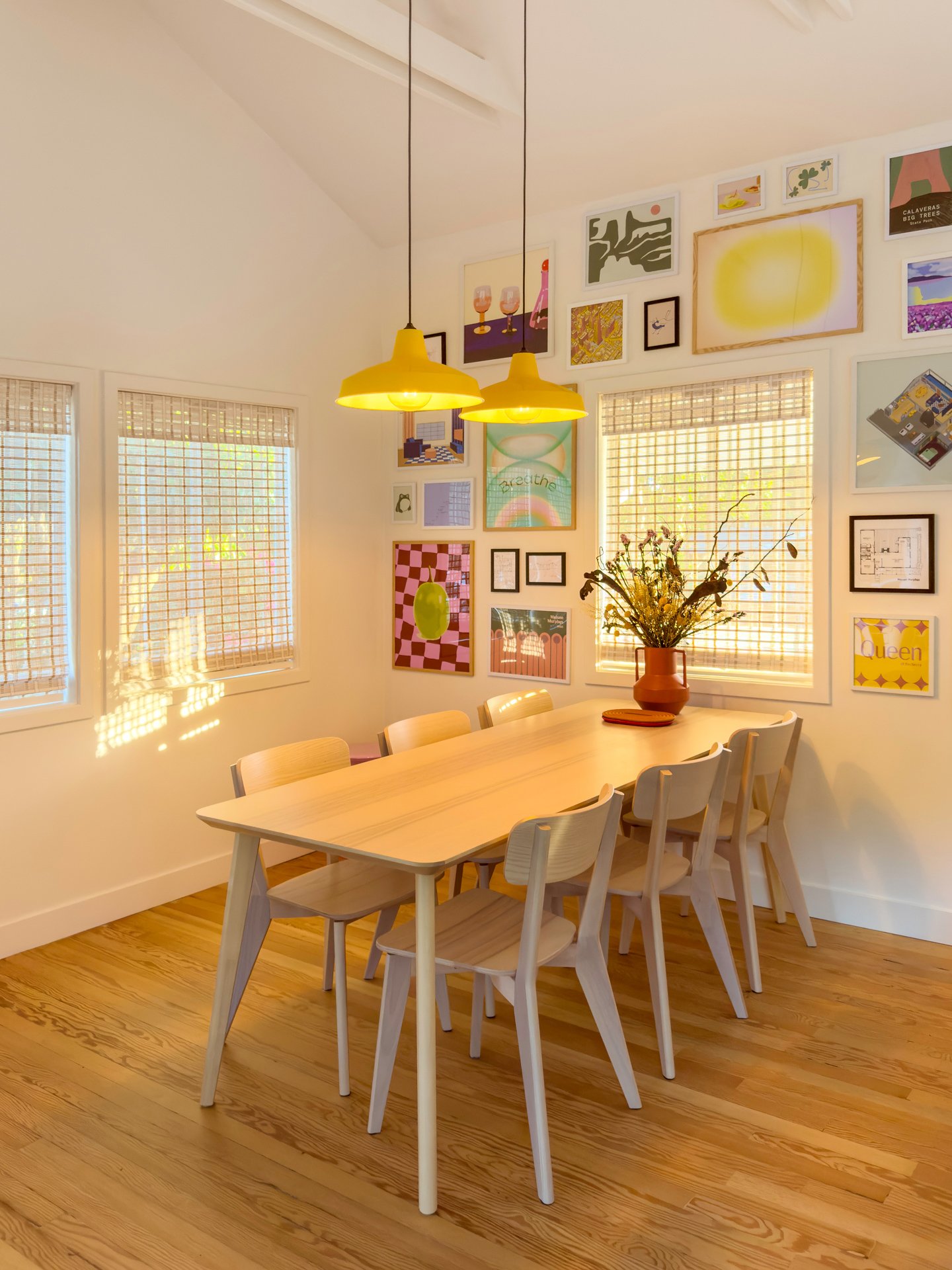 A dining room with a light wooden table, six matching chairs, and a large flower arrangement in a brown vase. The room features wooden flooring, multiple framed artworks on the wall, two yellow pendant lamps hanging from the ceiling, and two windows with blinds allowing sunlight to cast shadows inside.