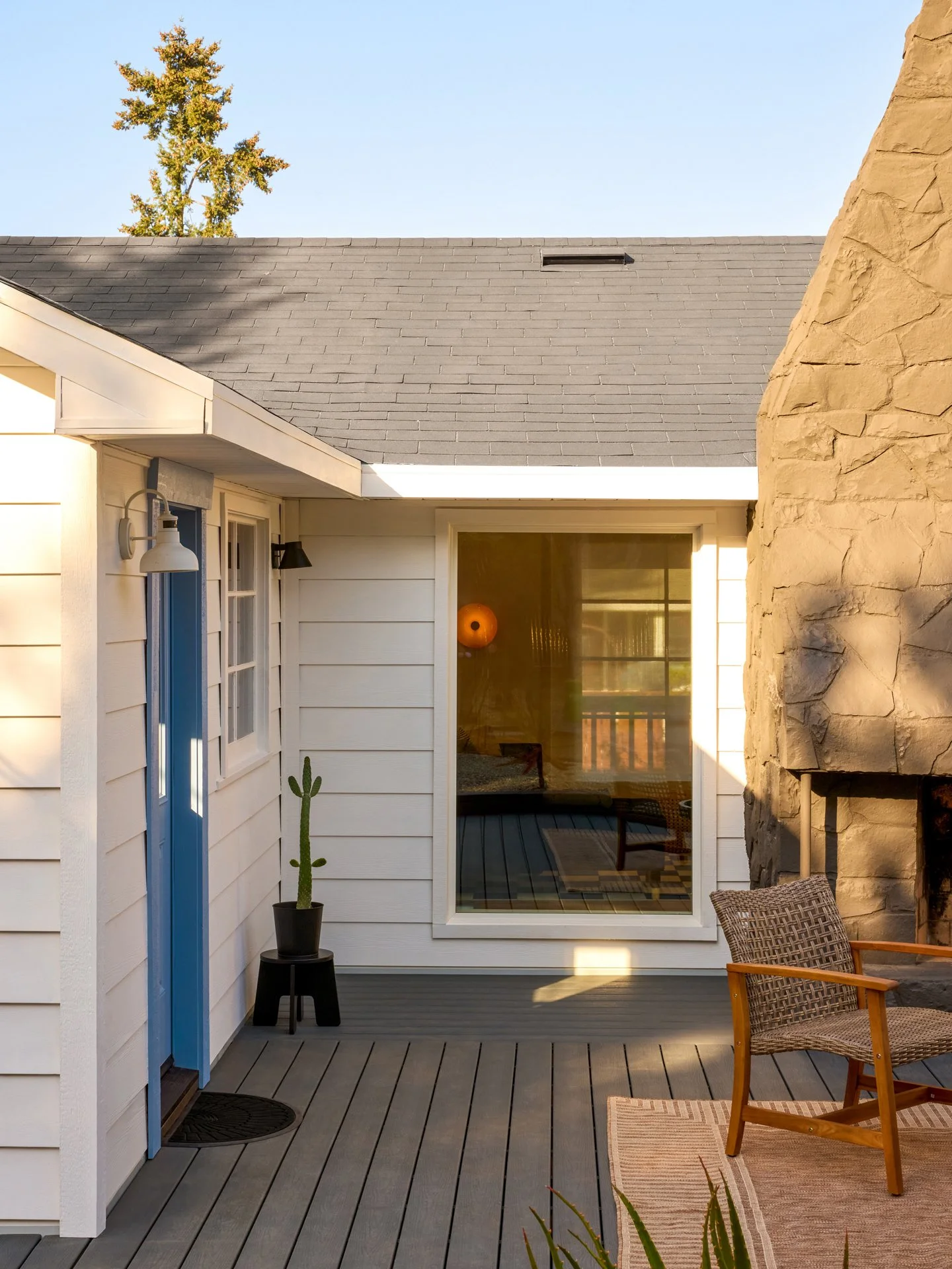 A wooden deck outside a house with a stone fireplace, a wicker chair, a small cactus in a black pot, and a sliding glass door showing an interior with a hanging orange light.