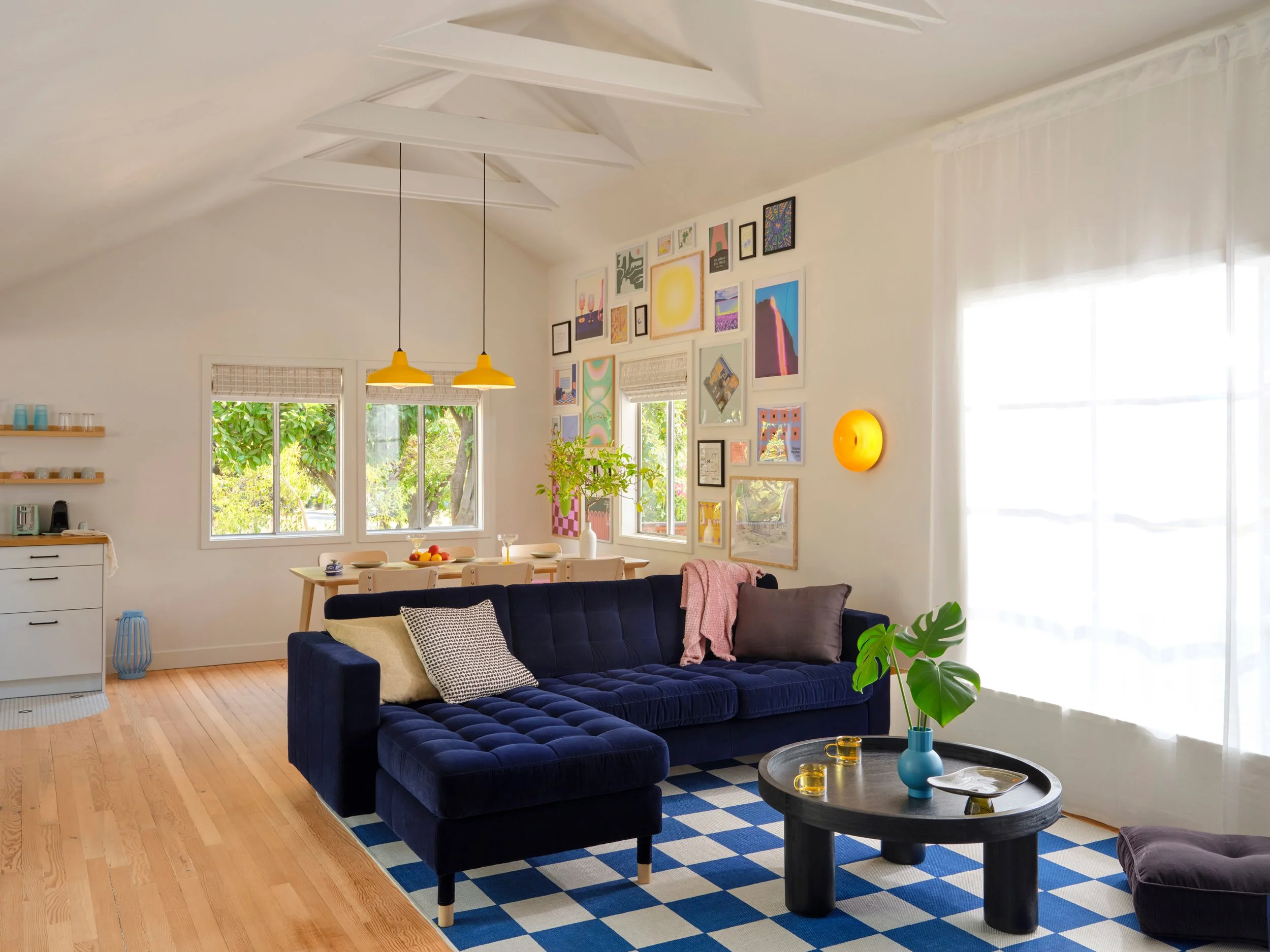 A living room with white walls and ceiling, a large window with curtains, a dark blue tufted sofa with pillows, a black coffee table with yellow candles and a blue vase with a green leafy plant, framed artwork on the walls, a wooden dining table with chairs, a yellow hanging lamp, and a blue and white checkered rug on a wood floor.