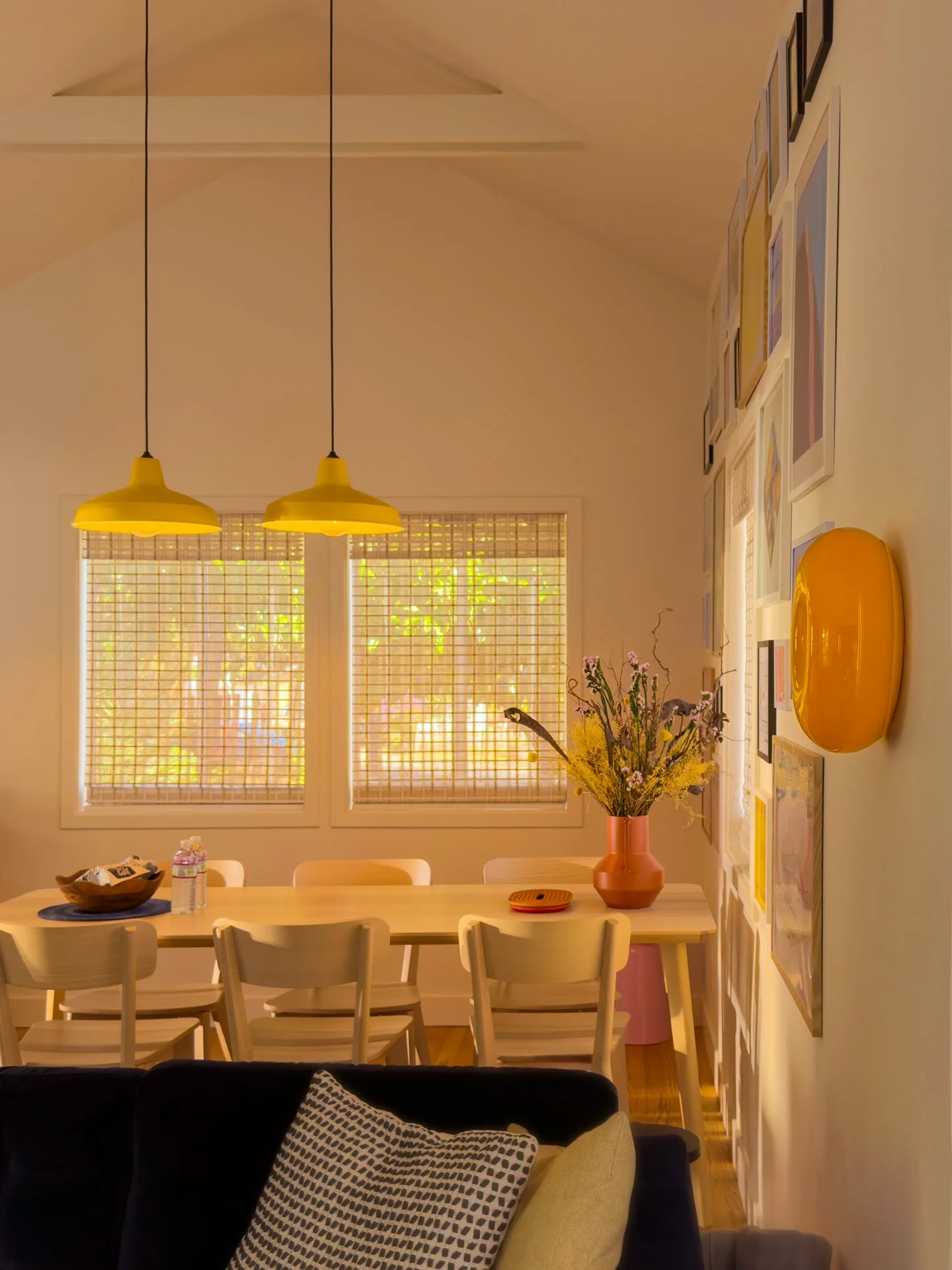 A dining room with a wooden table, six white chairs, and a large orange vase with flowers. Two yellow pendant lights hang above the table. A gallery wall with framed artwork is on the right, and sunlight filters through bamboo window shades.