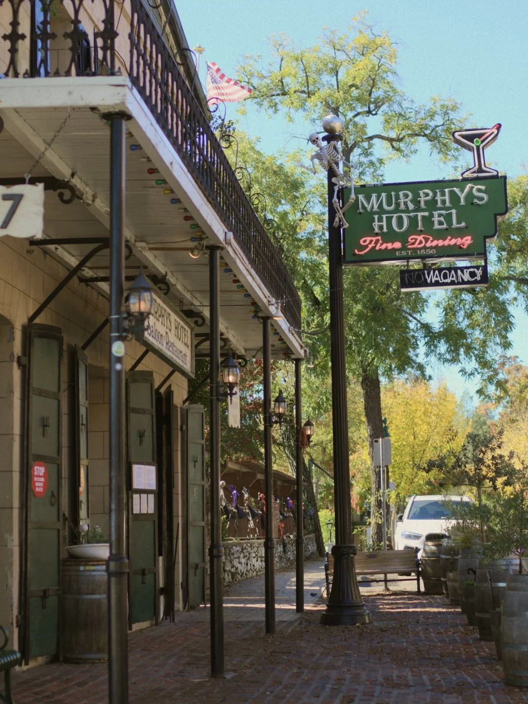 The exterior of Murphy's Hotel, a historic hotel established in 1856 with a sign indicating it is a fine dining establishment. The sign features a skeleton and a martini glass and says 'No Vacancy.' The hotel has a porch with hanging lanterns and outdoor seating, with a tree in the background and a parked white vehicle on the street.