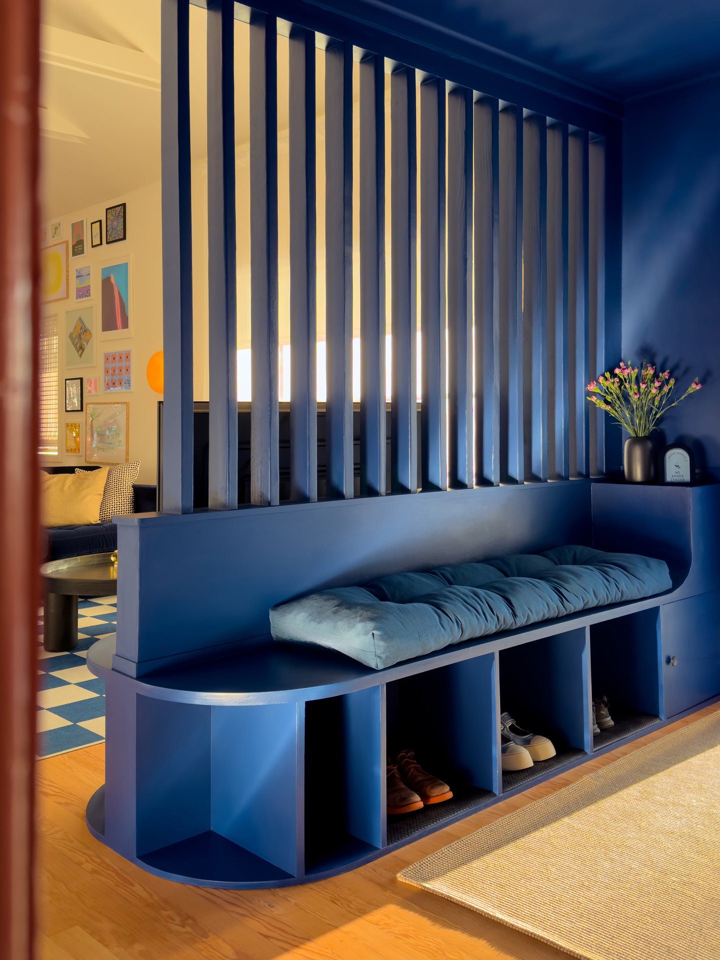 Blue built-in shelf with a cushioned seating area above shoe storage, a window with sunlight, a vase with pink flowers, and a dark blue wall.