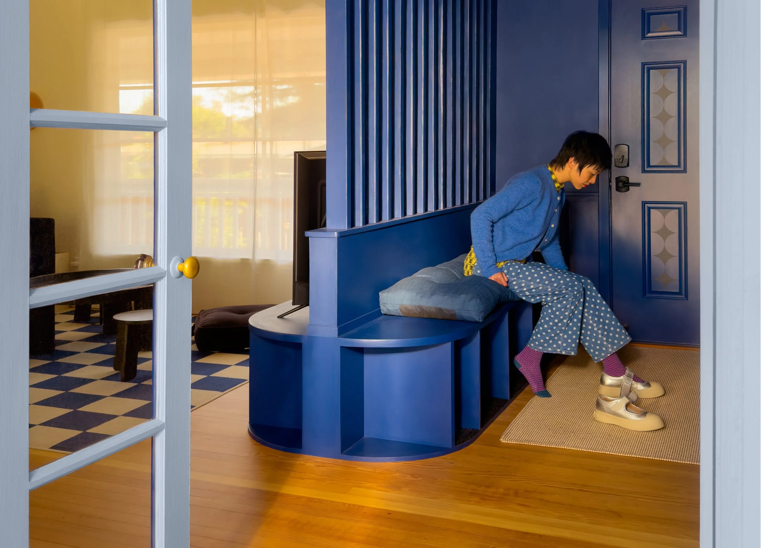 A woman with short dark hair sits on a blue built-in bench in a room with wooden flooring and a blue wall with decorative panels. She is wearing a blue jacket, polka-dotted pants, and gold shoes. Her slippers are on the floor nearby. There is a small cushion on the bench and a flat-screen TV in the background. The room is well-lit with natural light coming through a window.
