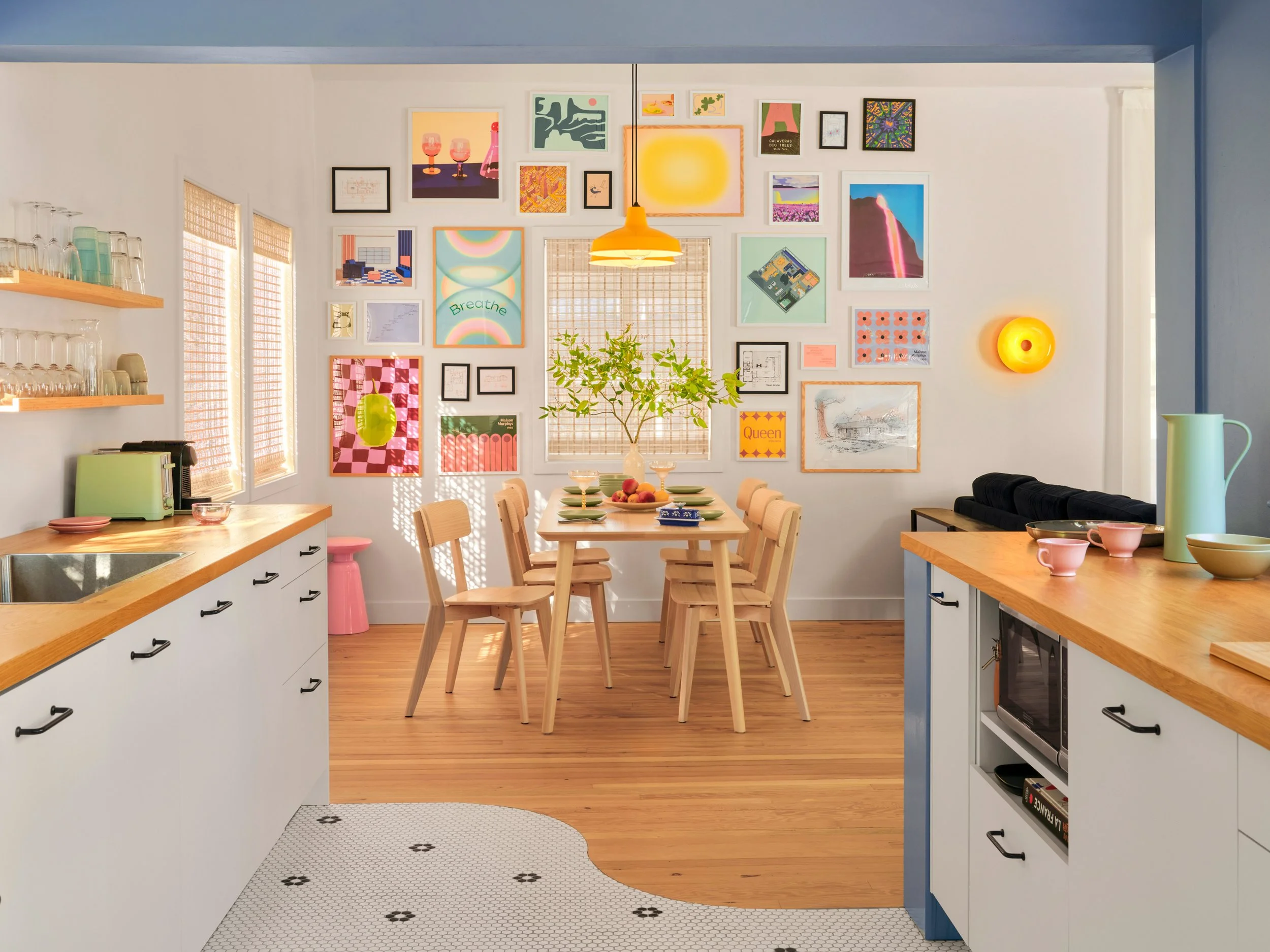 A kitchen and dining area with a gallery wall of colorful artwork and photos. The room features a wooden dining table with chairs, a hanging yellow light, and shelves with glassware and kitchen items. There is natural light coming through woven blinds.