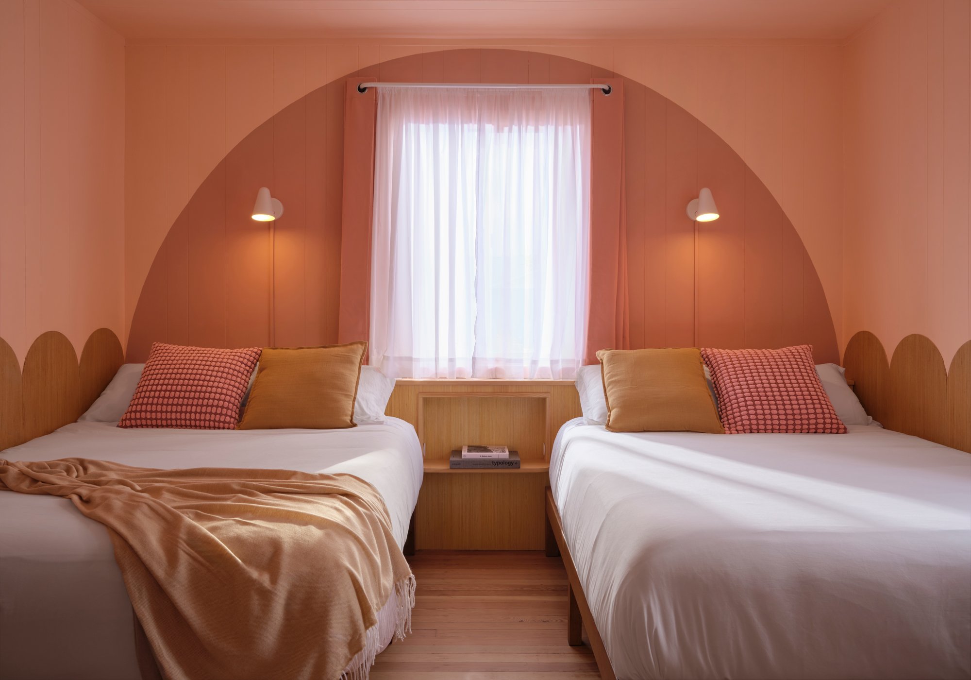 Two single beds with beige and patterned pillows, a shared wooden nightstand, a window with sheer white curtains, pink walls, and modern wall-mounted lamps.