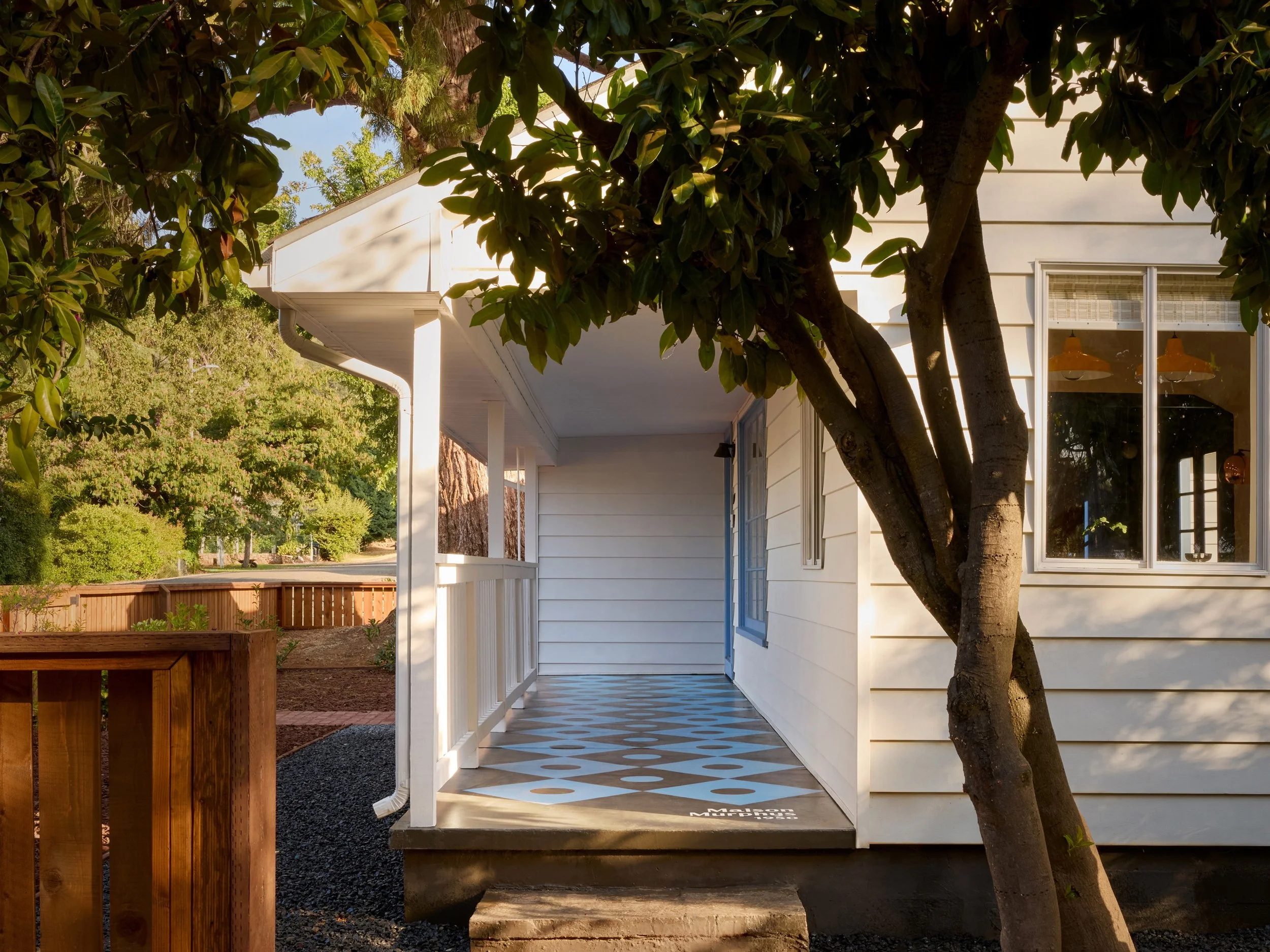 Front porch of a white house with a large tree partially blocking the view. The porch has a checkered black and white tile floor and a white railing. A blue door is visible. Sunlight filters through the tree leaves.