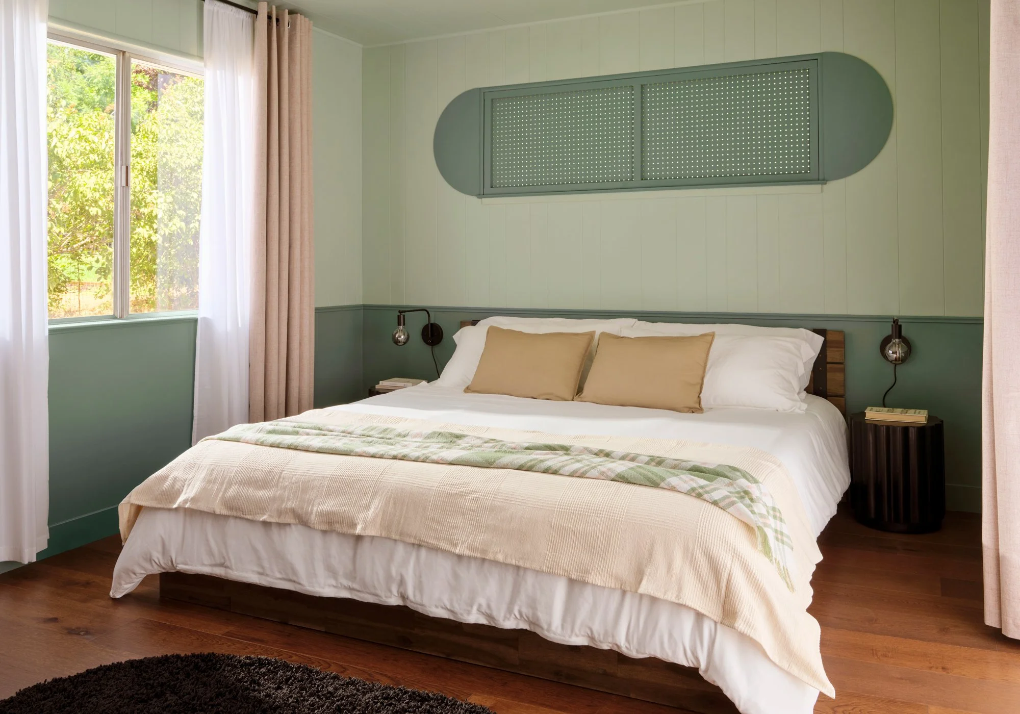 A cozy bedroom with a large bed, beige and white bedding, and two beige pillows. A black round side table with a book is next to the bed. Behind the bed, a green wall with a rectangular, perforated window and wood paneling. A window with white and be