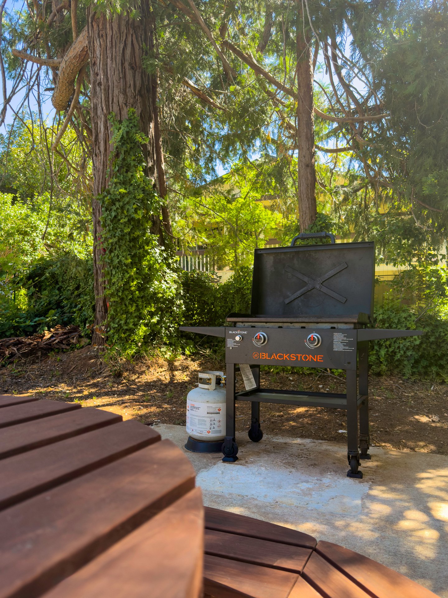 A Blackstone gas grill with lid open is outdoors on a patio, with a propane tank attached underneath. The background features trees and greenery, and there's a wooden table in the foreground.
