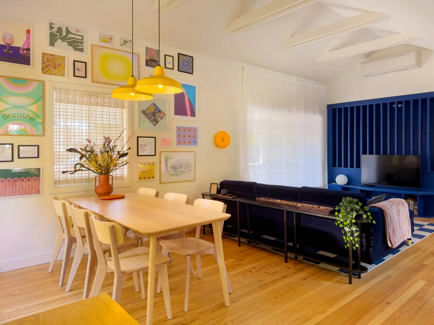 A dining room with a wooden table and cream-colored chairs, a gallery wall with various colorful pictures, a window with blinds, a yellow pendant light, a blue built-in media unit, and decorative plants.