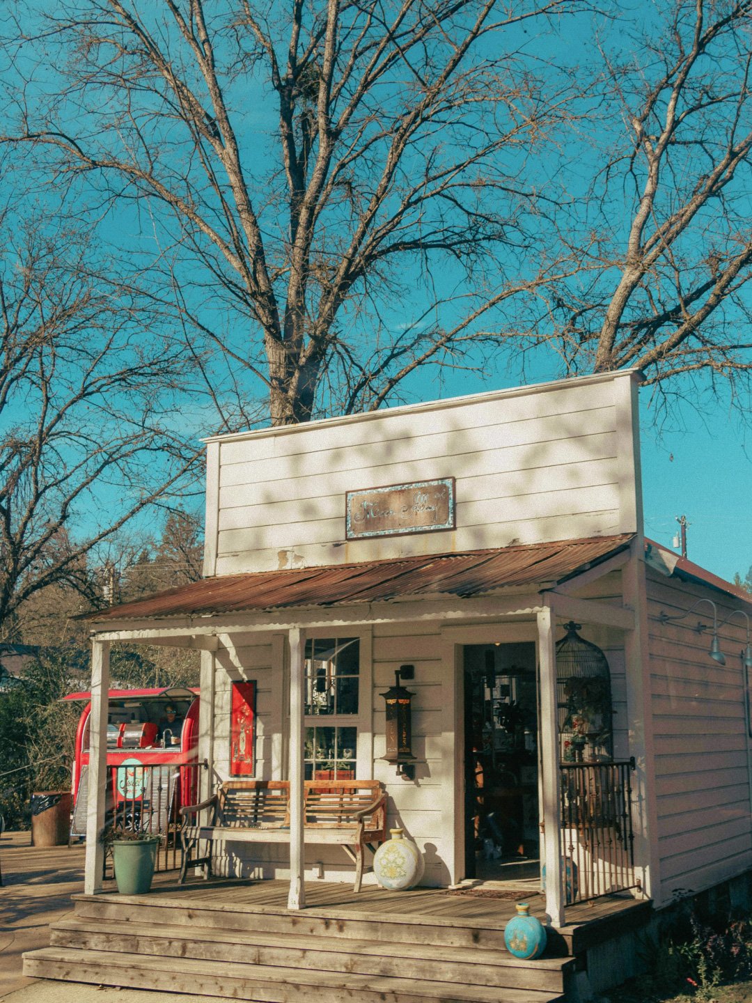 Small white wooden shop with a rusty metal roof, a porch with benches, and decorative items outside, set against a backdrop of leafless trees and a blue sky.