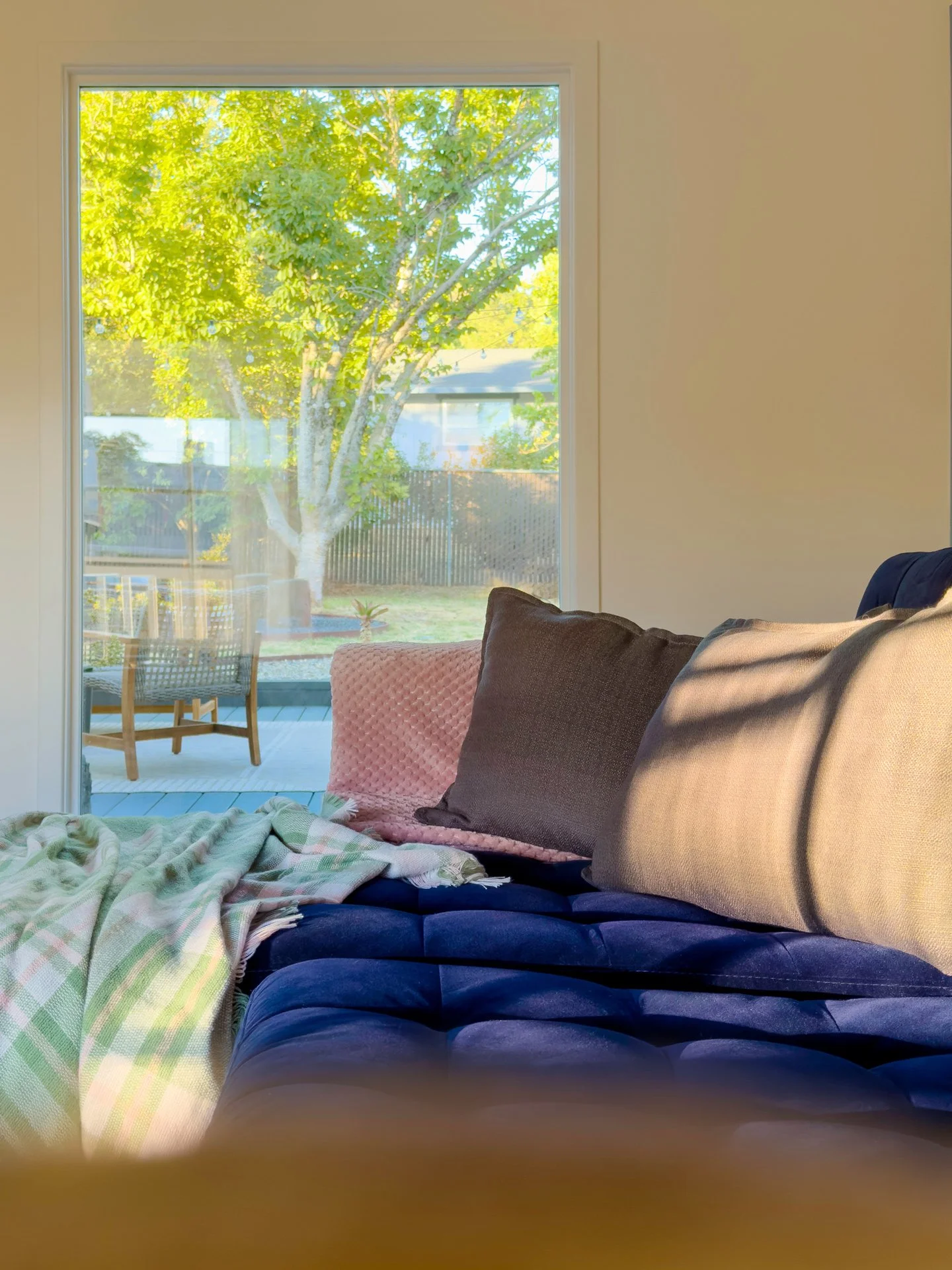 View of a living room with a beige wall and a large window showing a tree with green leaves outside. Inside, a blue sofa with various pillows and a blanket is visible in the foreground. Outside, there is a patio with chairs and a fence.