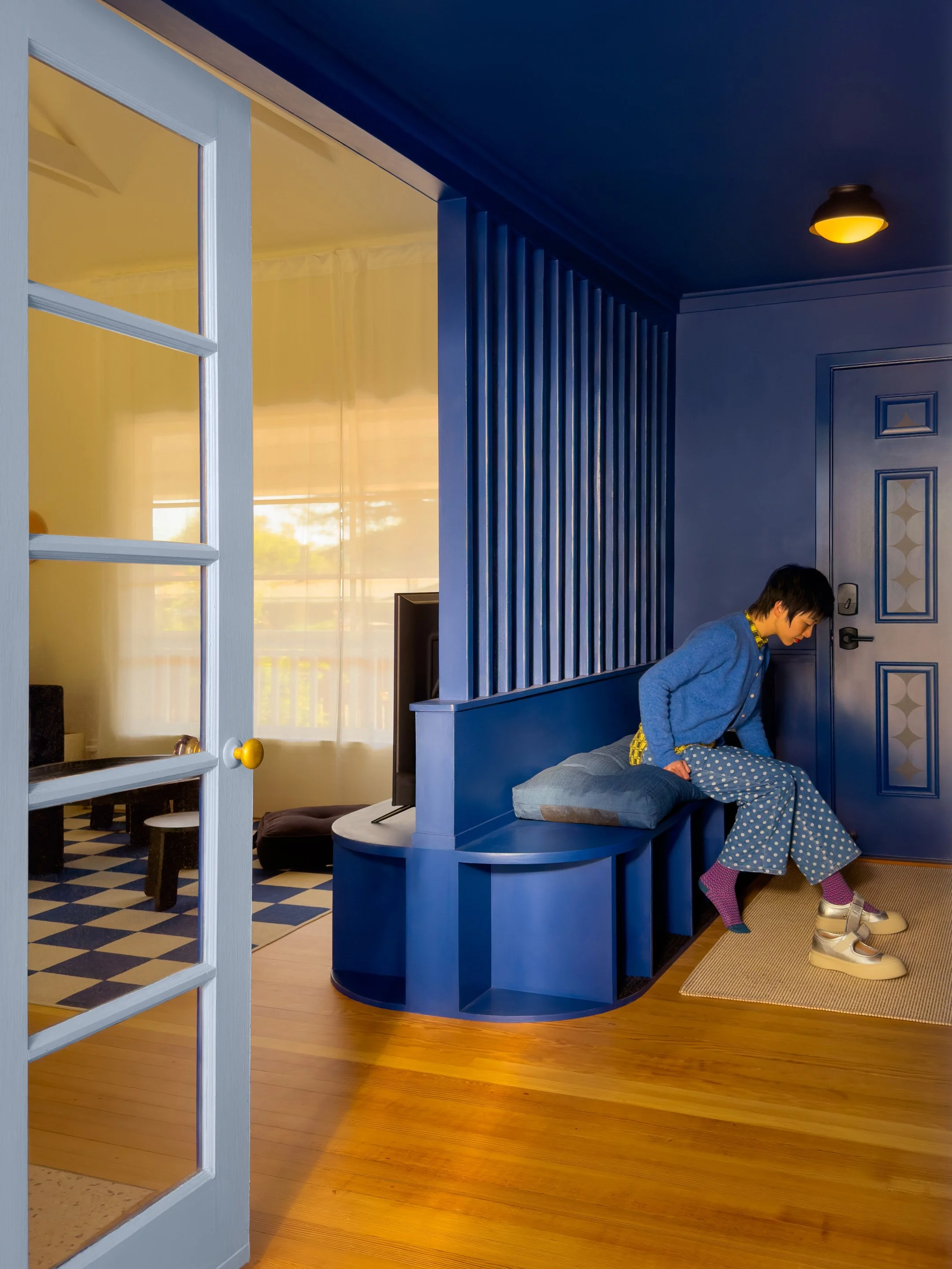 A person with dark hair wearing a blue sweater and patterned pajama pants sitting on a built-in blue bench next to a door, with a yellow patterned shirt underneath and white sneakers with purple socks, inside a room with wooden floors, blue walls, and a ceiling lamp.