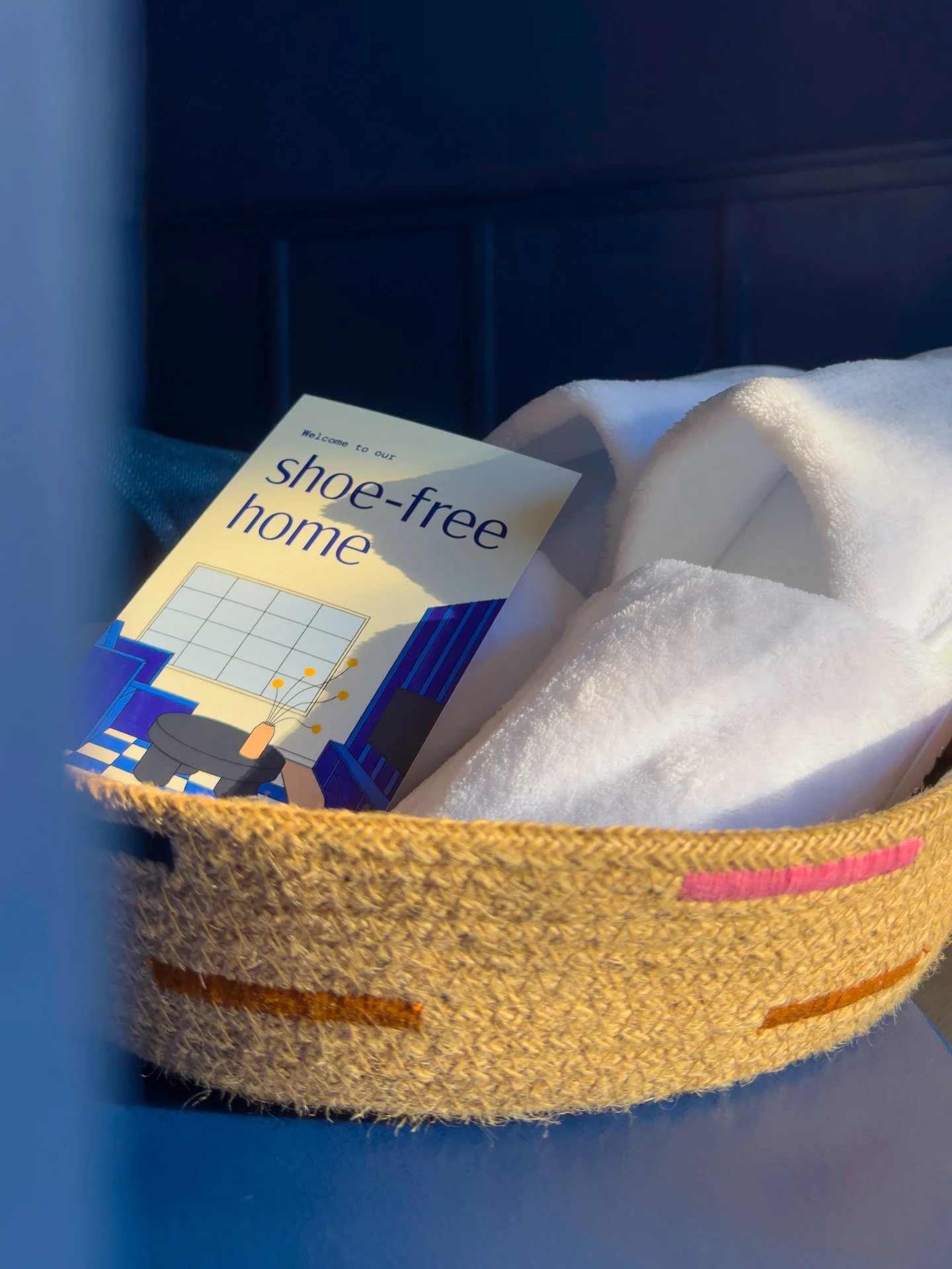 Close-up of a beige woven bag with pink stitching, containing a shoe-free home brochure and a white towel, on a blue surface.