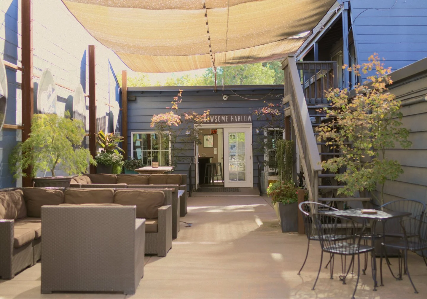 Outdoor patio area with beige cushioned sectional sofas, black metal chairs, potted plants, a small table, and a building with stairs and a sign that reads "The Awesome Harlow."