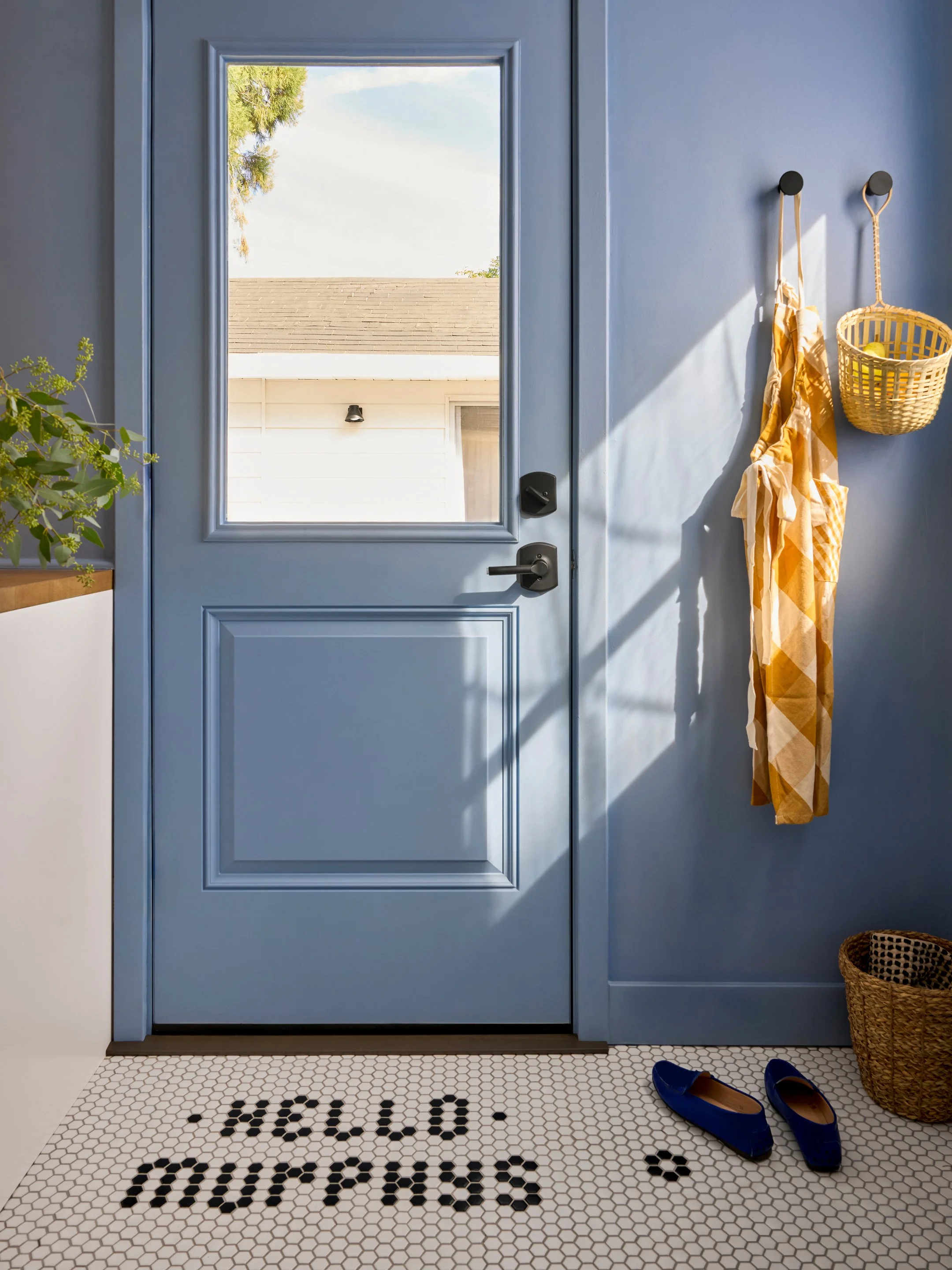 Blue door with glass window showing roof and trees outside, white hexagonal floor with black letters spelling 'HELLO' and 'GOODBYE', blue shoes, wicker basket, yellow checkered cloth hanging on wall hooks, and a plant on a table.