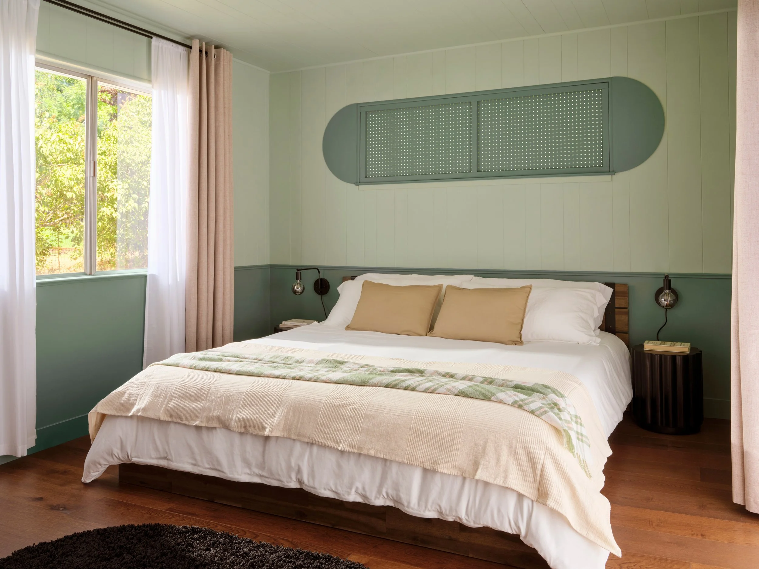 A cozy bedroom with a large bed featuring beige pillows and a striped blanket, dark wood flooring, a window with white and beige curtains, green walls, a black round side table, and modern black wall-mounted lamps.