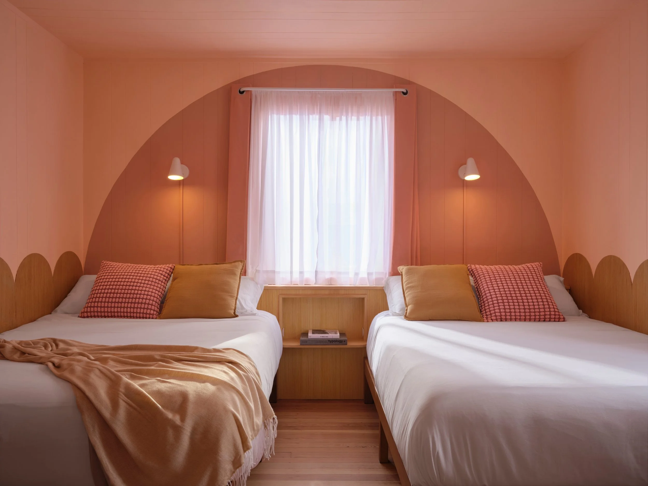 A bedroom with two beds, each with white bedding and multiple pillows, pink and beige throw blankets, a window with sheer white curtains, and two wall-mounted lamps on either side of the window.