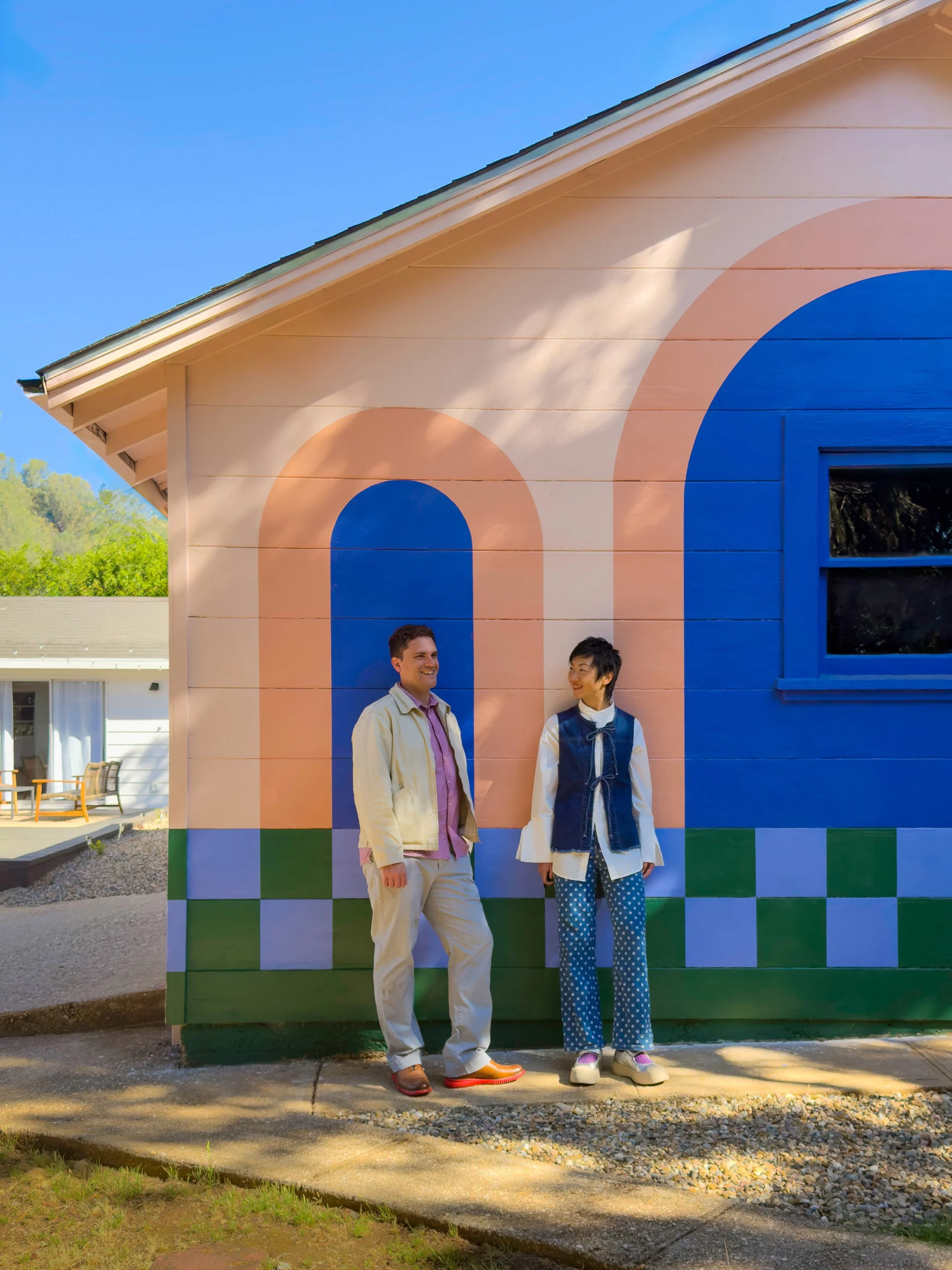 Two people standing in front of a colorful building with geometric painted designs, smiling and looking at each other on a sunny day.