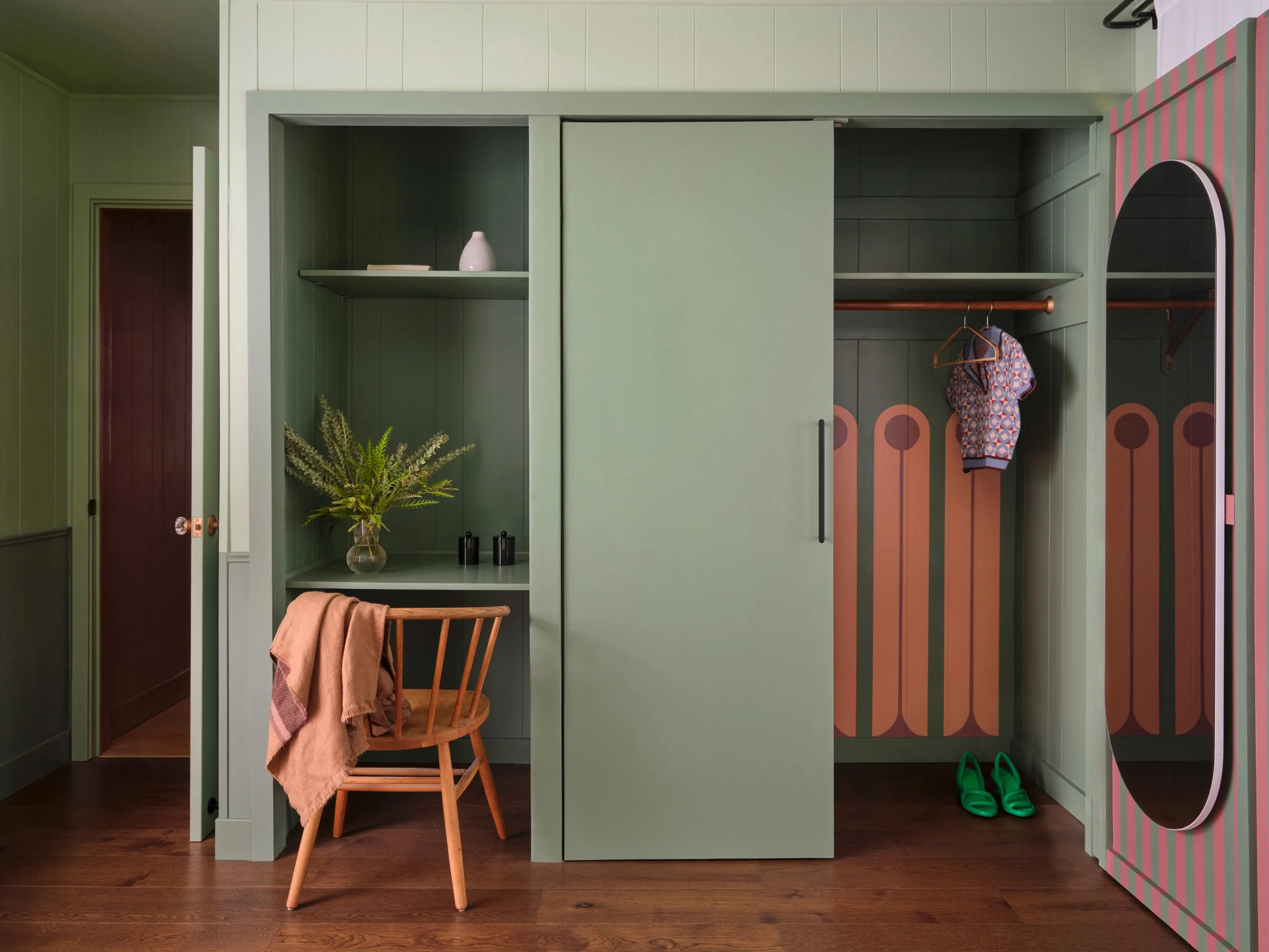 A room with green built-in wardrobe and a small wooden chair. The wardrobe has open shelves and a hanging rod with a patterned shirt. A pink vase and books are on the upper shelf. There is a plant on the chair and green slippers on the floor.