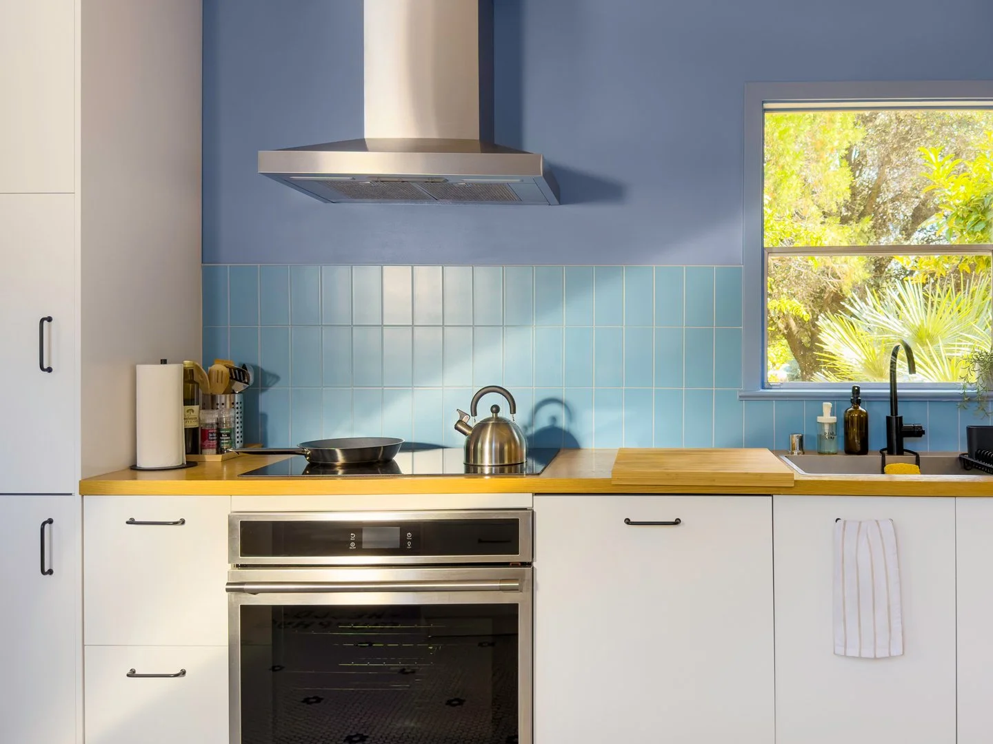 A modern kitchen with white lower cabinets, a light wood countertop, and a stainless steel oven. There is a stainless steel range hood above the stovetop and a large window to the right, showing trees and greenery outside. Blue tiles are on the wall behind the counter.