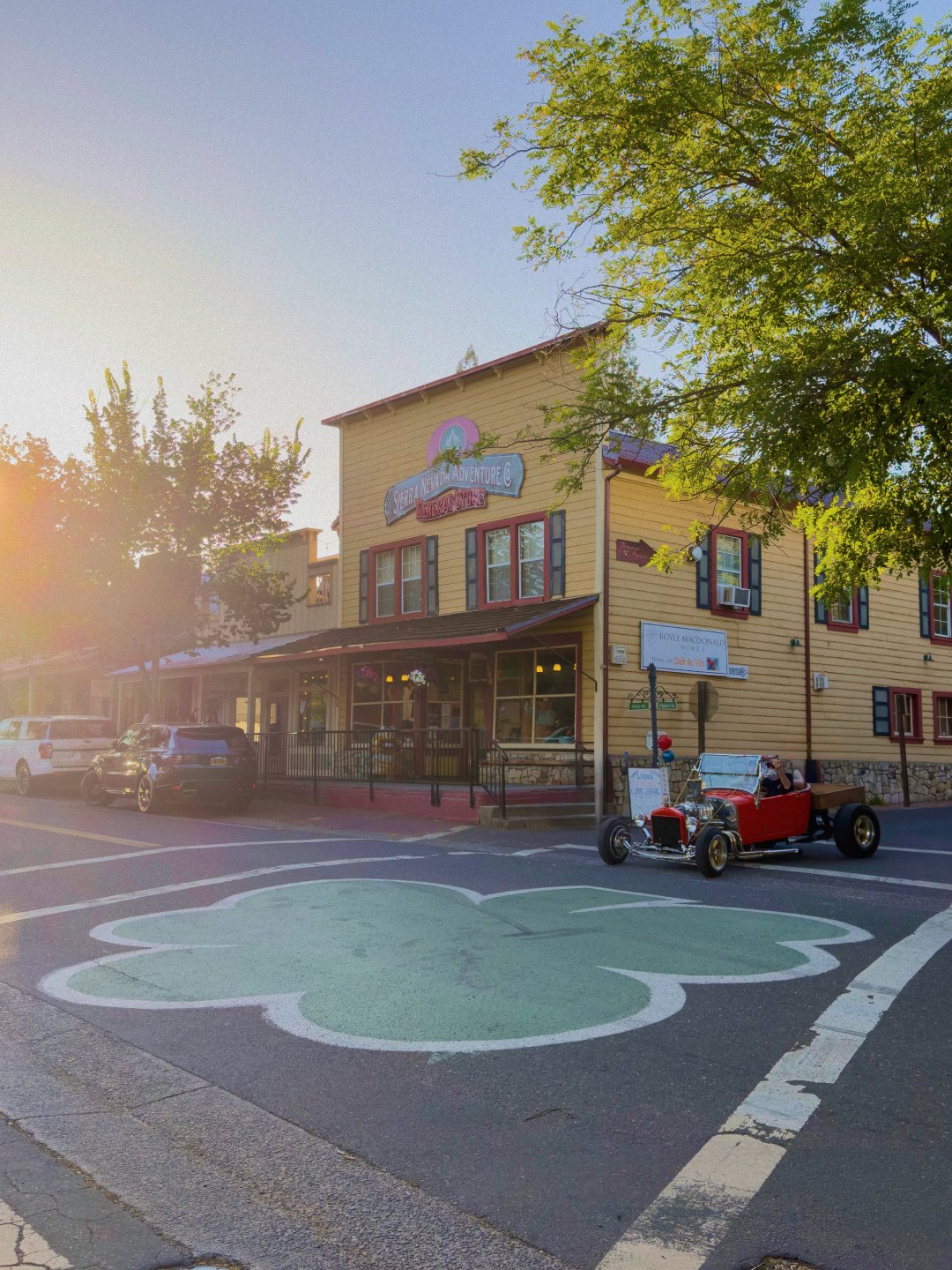 A street scene in front of a yellow building with a sign that reads 'Sierra Nevada Adventure & Dispensary Store.' A vintage red car is parked on the street, near a large shamrock-shaped painted area on the road. There are trees, a few parked cars, and the sun setting in the background, casting a warm glow.
