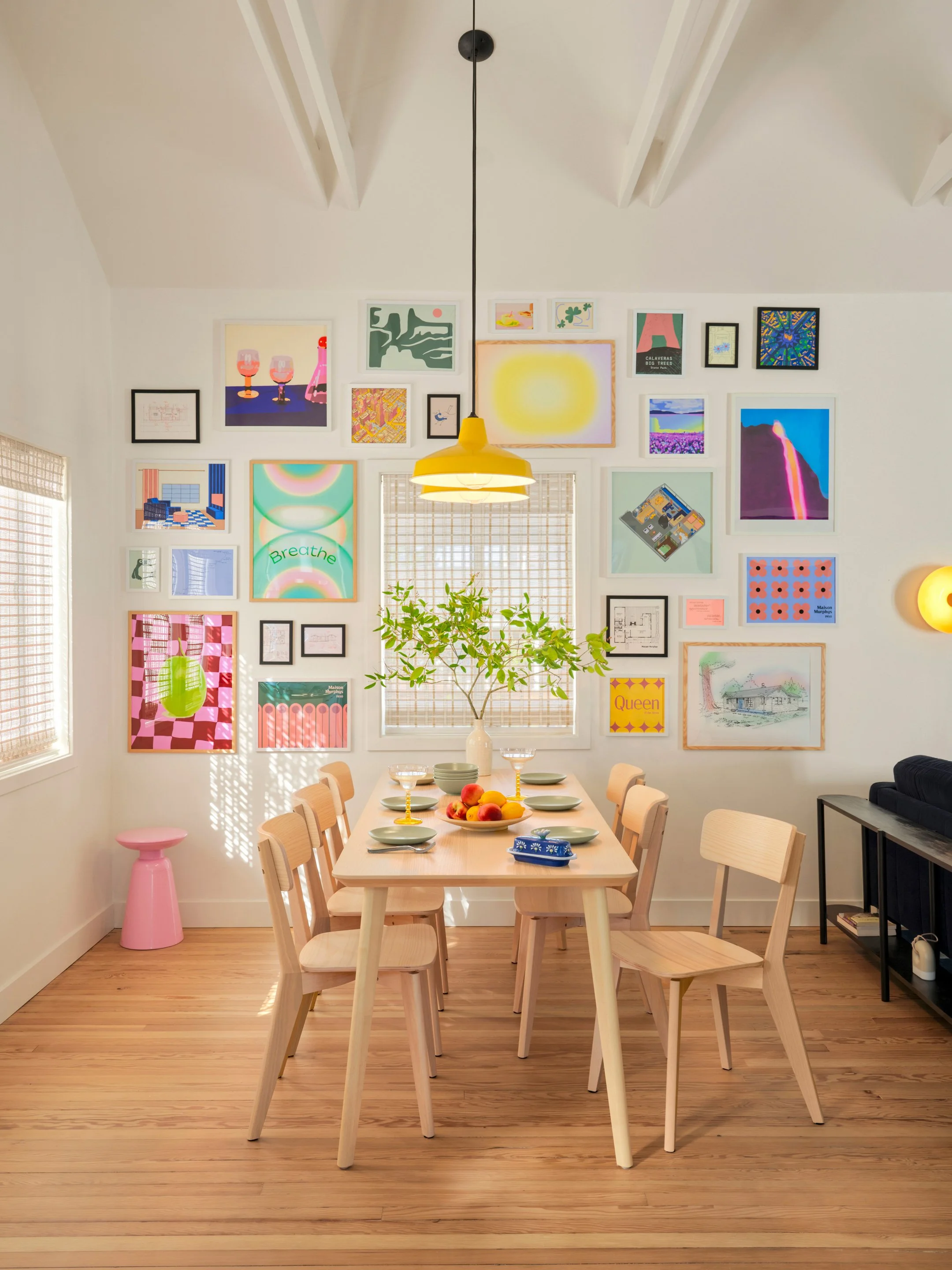 A dining room with a long light wood table, surrounded by matching chairs. The table is set with bowls, a plate of peaches, and yellow candleholders. The room features a gallery wall with colorful artwork and a window with blinds. A green plant in a white vase is on the table. A yellow hanging light fixture provides illumination.