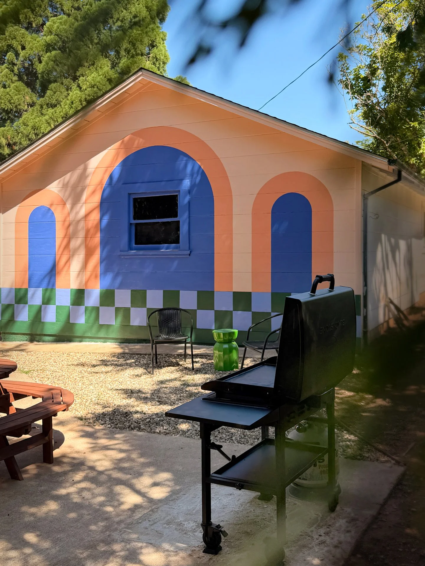 A house with colorful geometric mural art on the exterior wall, featuring blue, orange, and green shapes, with outdoor chairs, a barbecue grill, and a green planter in the yard, and trees in the background.