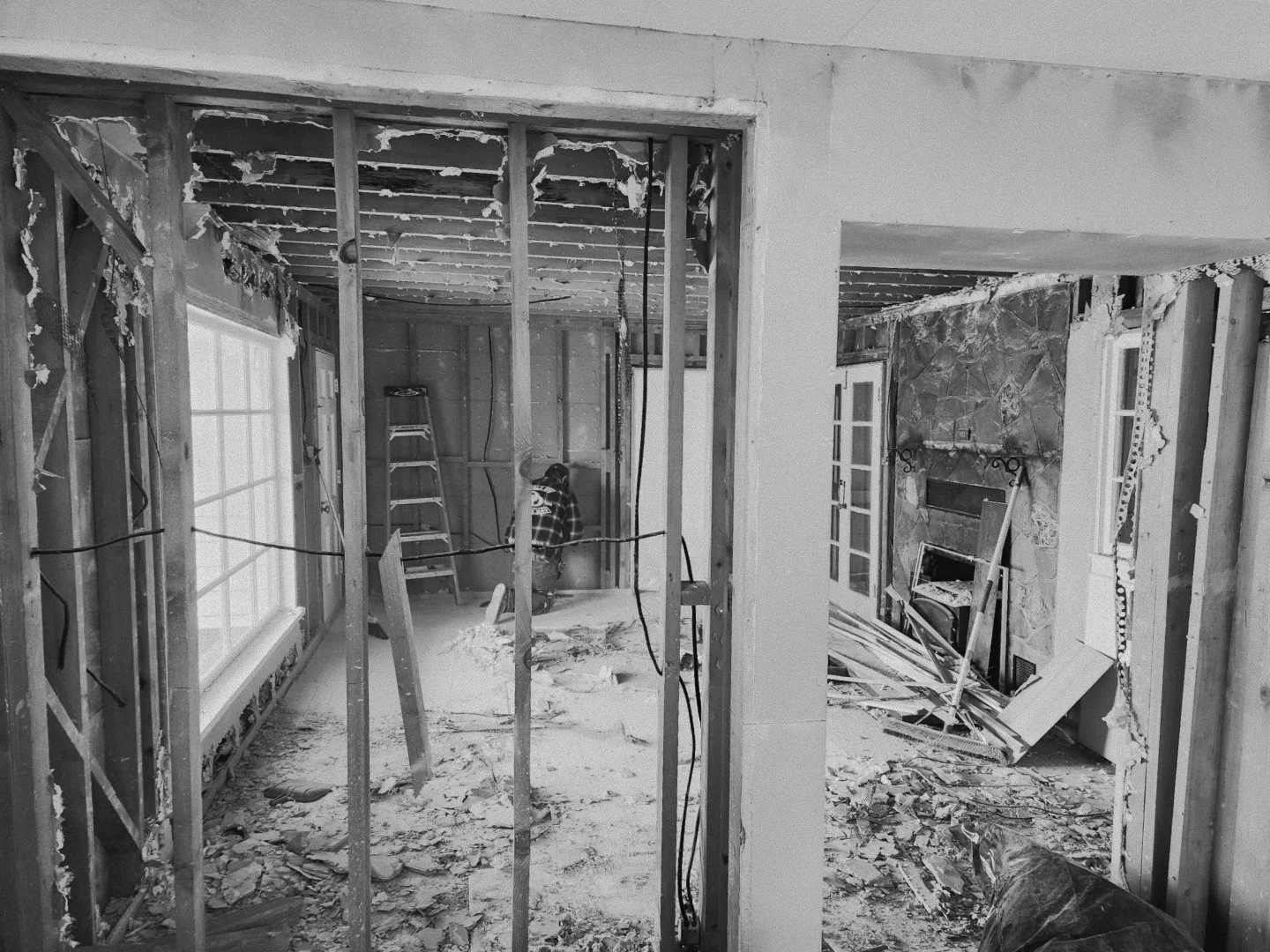 Interior of a house under renovation with exposed wall studs, debris on the floor, and a person in a plaid shirt working in the background.