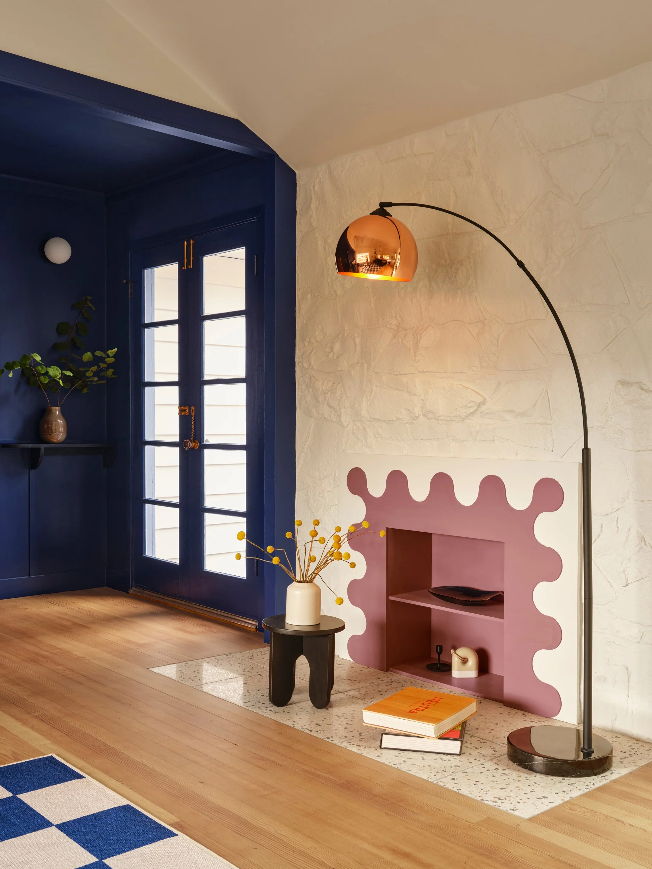 Interior of a room with a blue door, a pink scalloped wall shelf, a black side table with a white vase and yellow floral arrangement, a curved floor lamp with a reflective copper shade, and books on the floor. The wall has textured white stone, and the floor is hardwood with a small section of speckled terrazzo.
