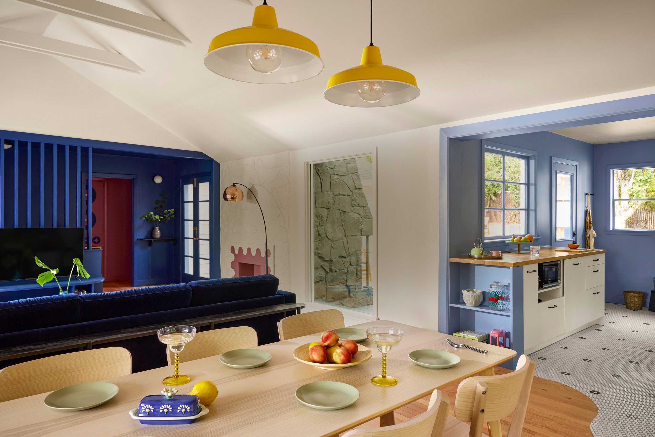 A dining area with a light wood table set with green plates, apples, and yellow candlestick holders with glasses. The background shows a blue wall with windows, a white kitchen with black handles, and a living room with a black sofa, blue shelving, and yellow pendant lights hanging from the ceiling.