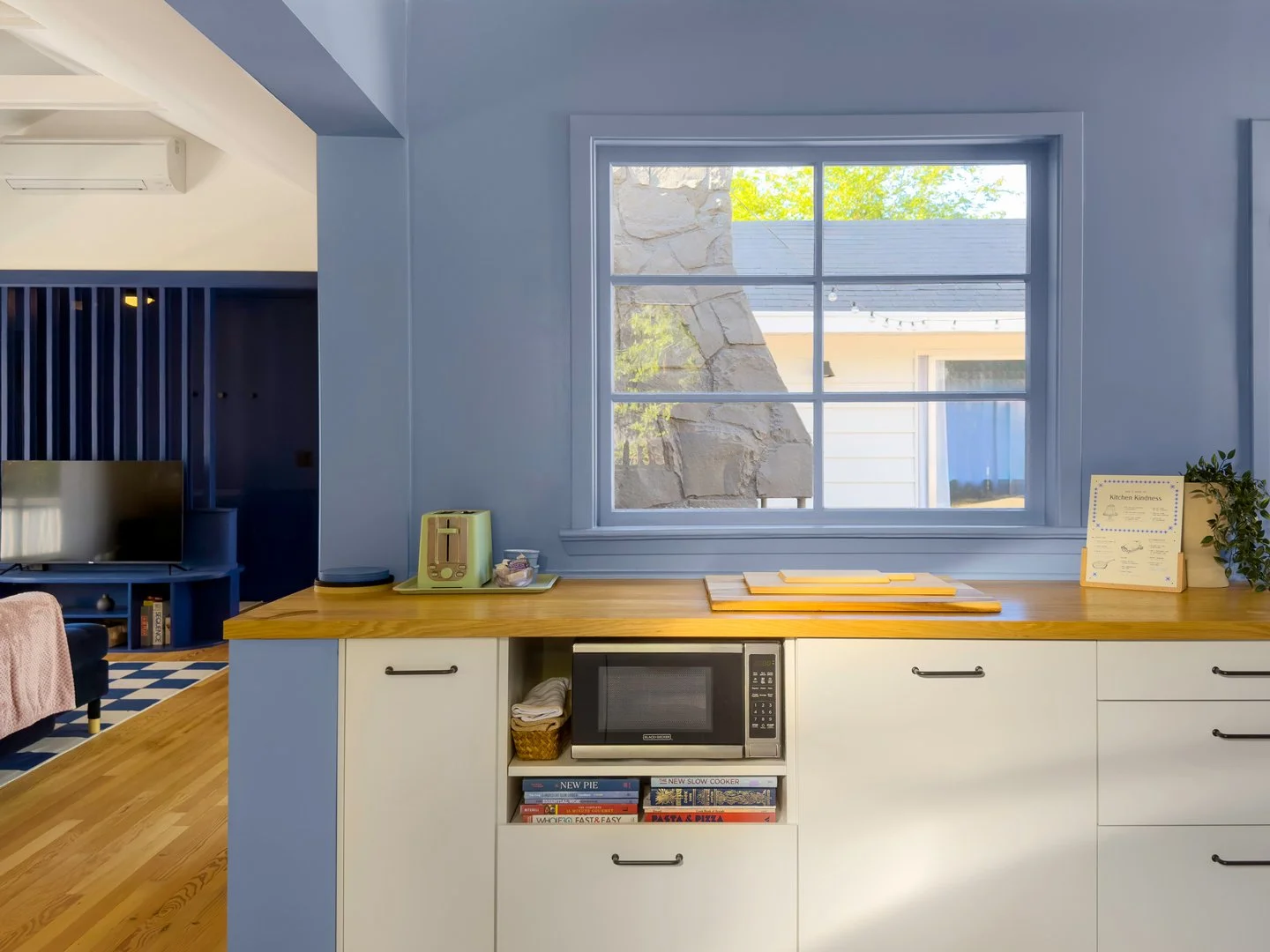 View of a light blue wall with a large window showing trees and a building outside, tall blue slatted partition, part of a living room with a TV and a small table.