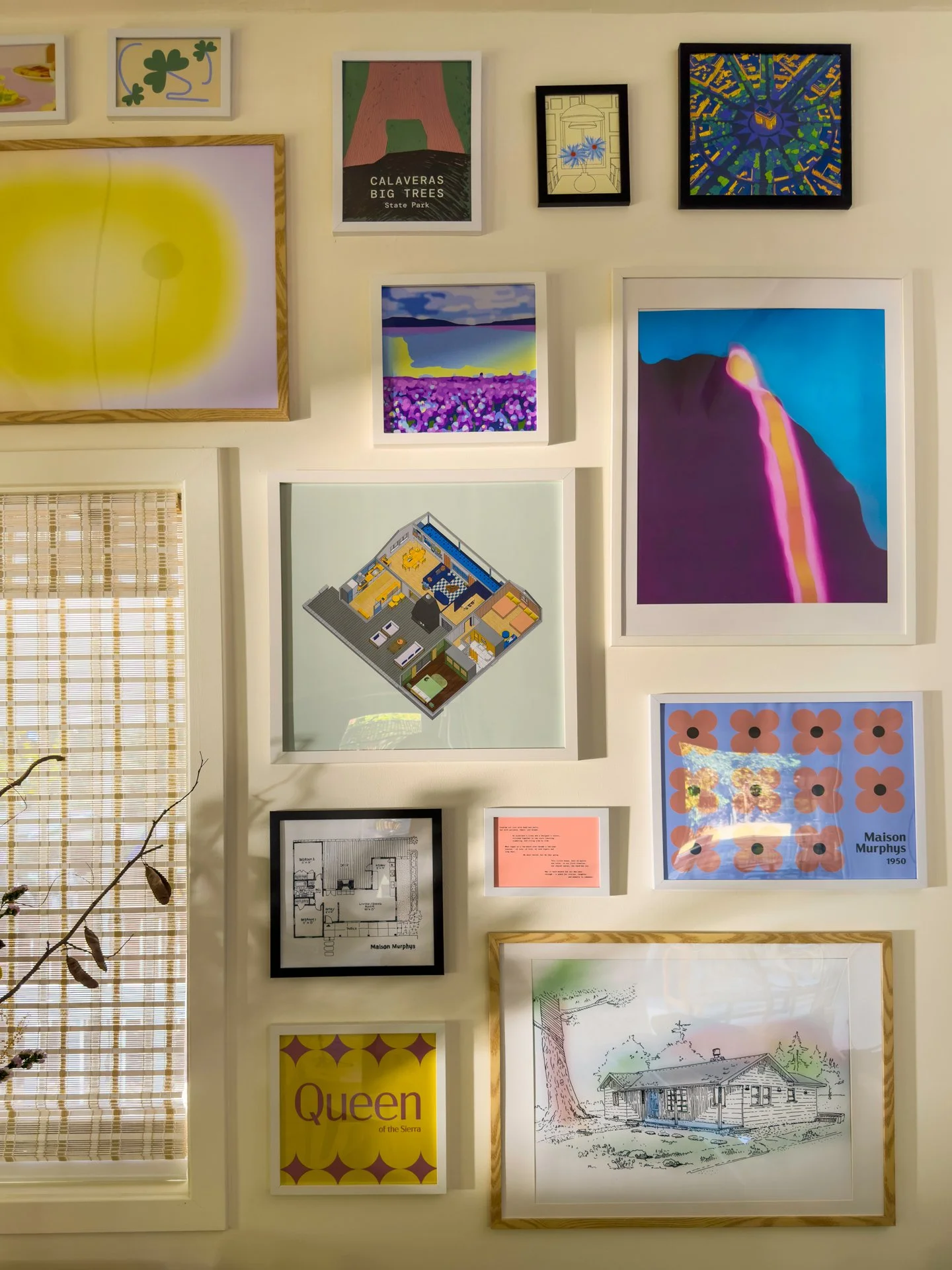 A gallery wall with various framed artworks, including colorful abstract art, landscape paintings, architectural blueprints, and a poster titled 'Queen of the Sierra.' The wall also features a window with bamboo blinds and a small branch with leaves in the foreground.