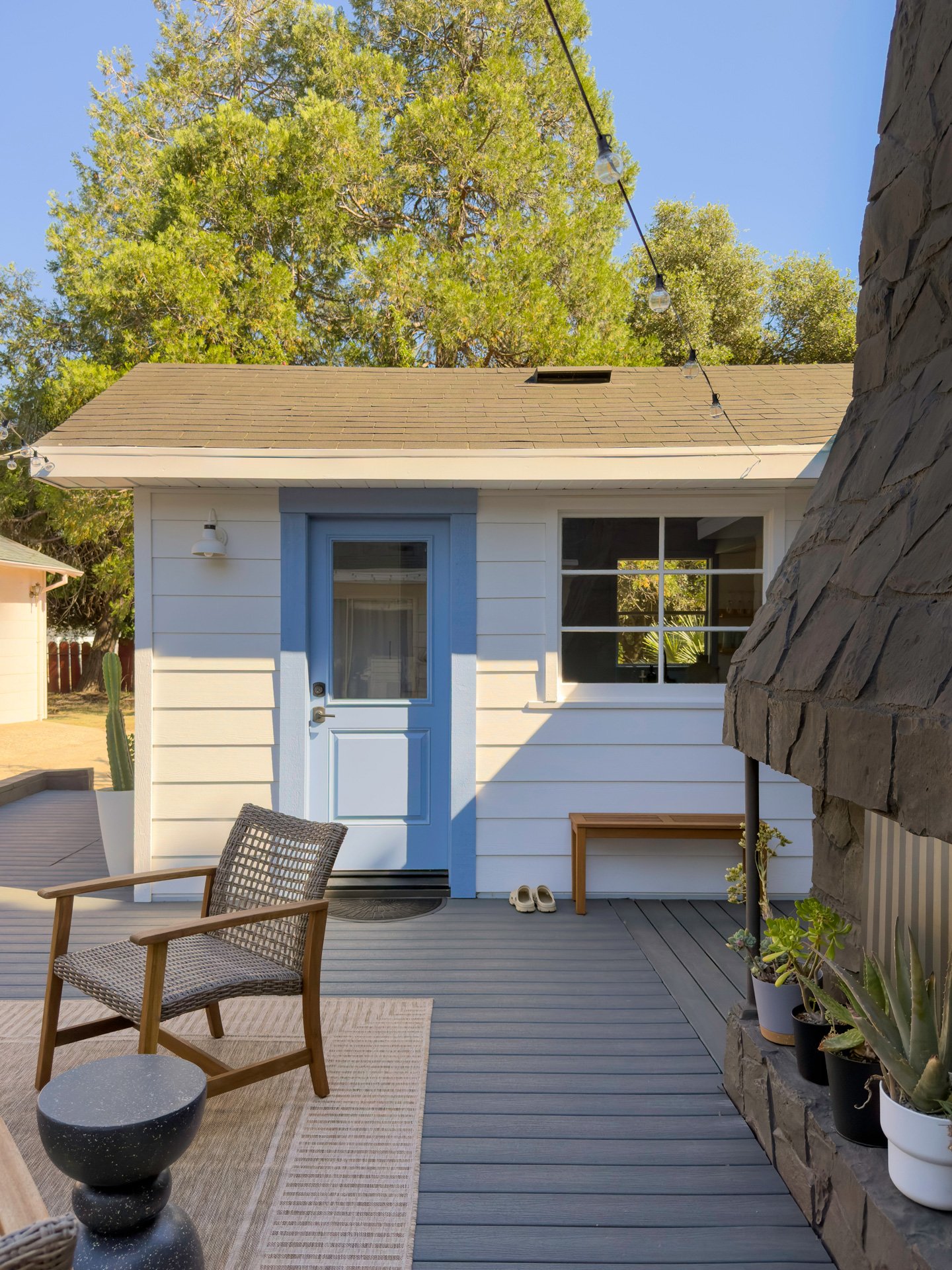 A backyard patio with outdoor furniture, potted plants, string lights, and the exterior of a white house with blue trim and a window.