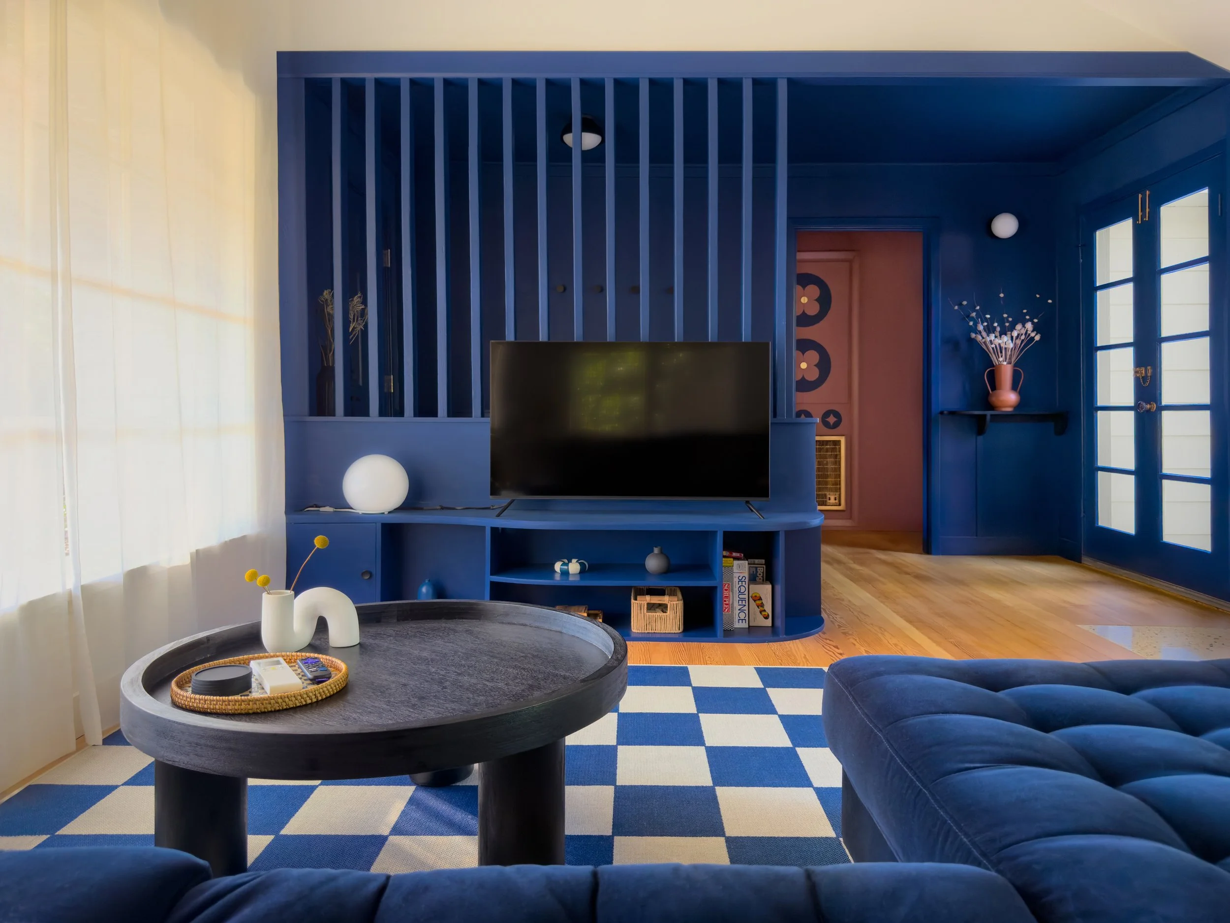Living room with blue and white checkered rug, dark blue velvet sofa, round black coffee table with white vases and a basket, blue wall with wooden slats, TV, books, and decorative items, sunlight coming through large windows.