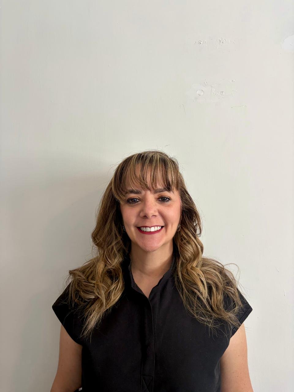 Mujer sonriendo con cabello rubio y ondulado, vestida con blusa negra, frente a una pared blanca