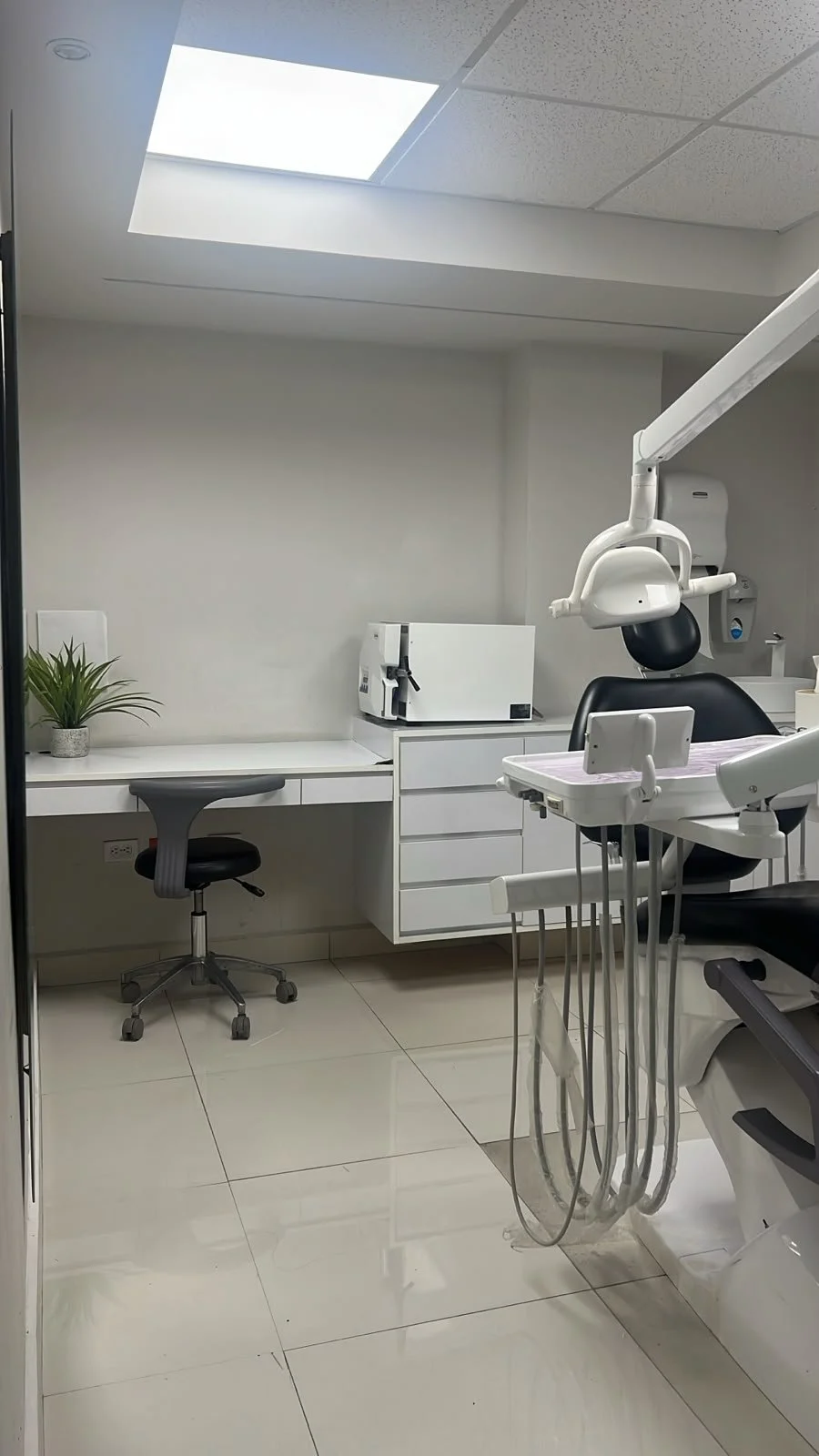 Modern dental treatment room with a dental chair, dental equipment, and a desk. Features a plant and natural light from the skylight above.