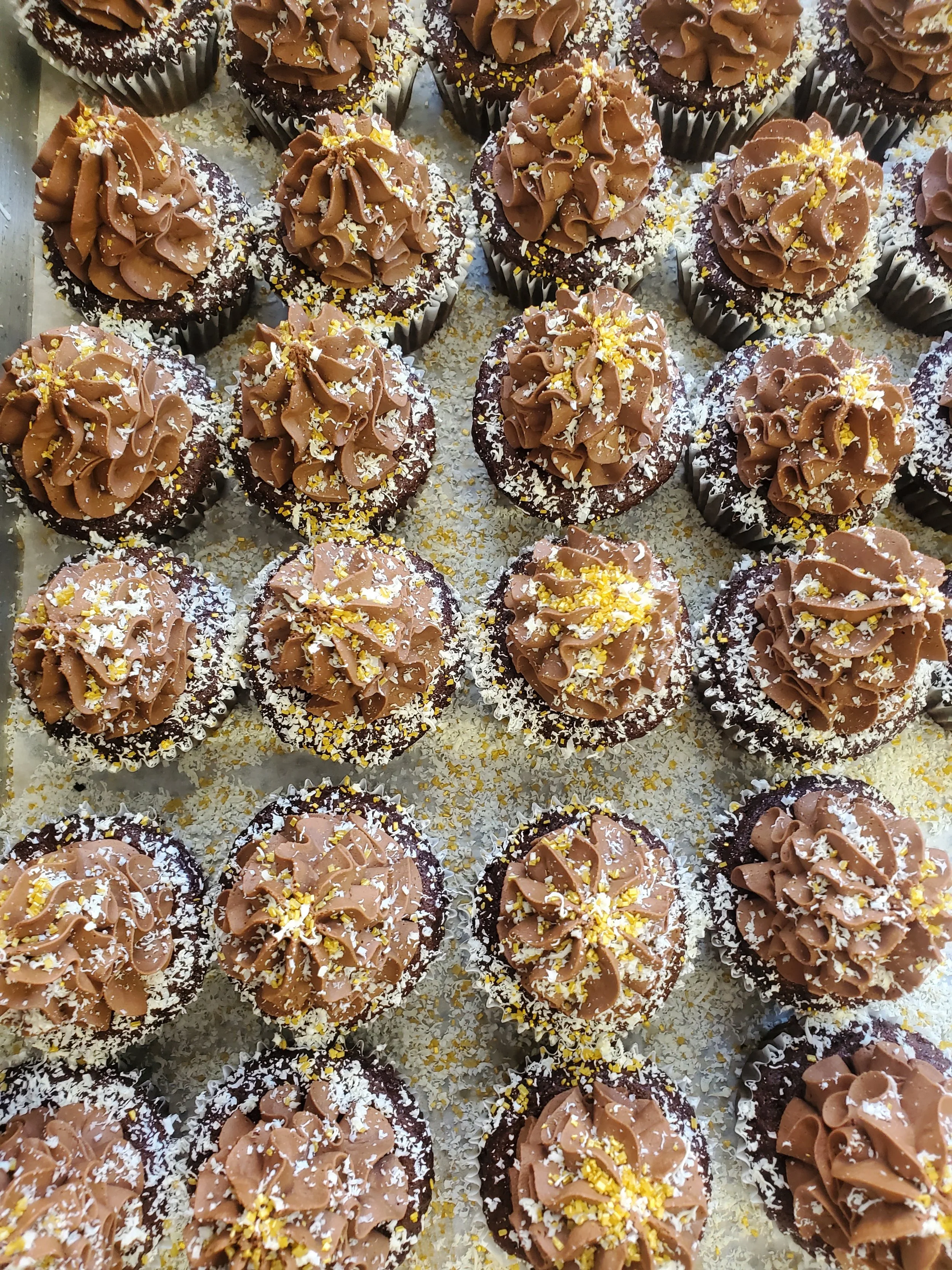 Close-up of multiple cupcakes with chocolate frosting, sprinkled with shredded coconut and yellow sprinkles.