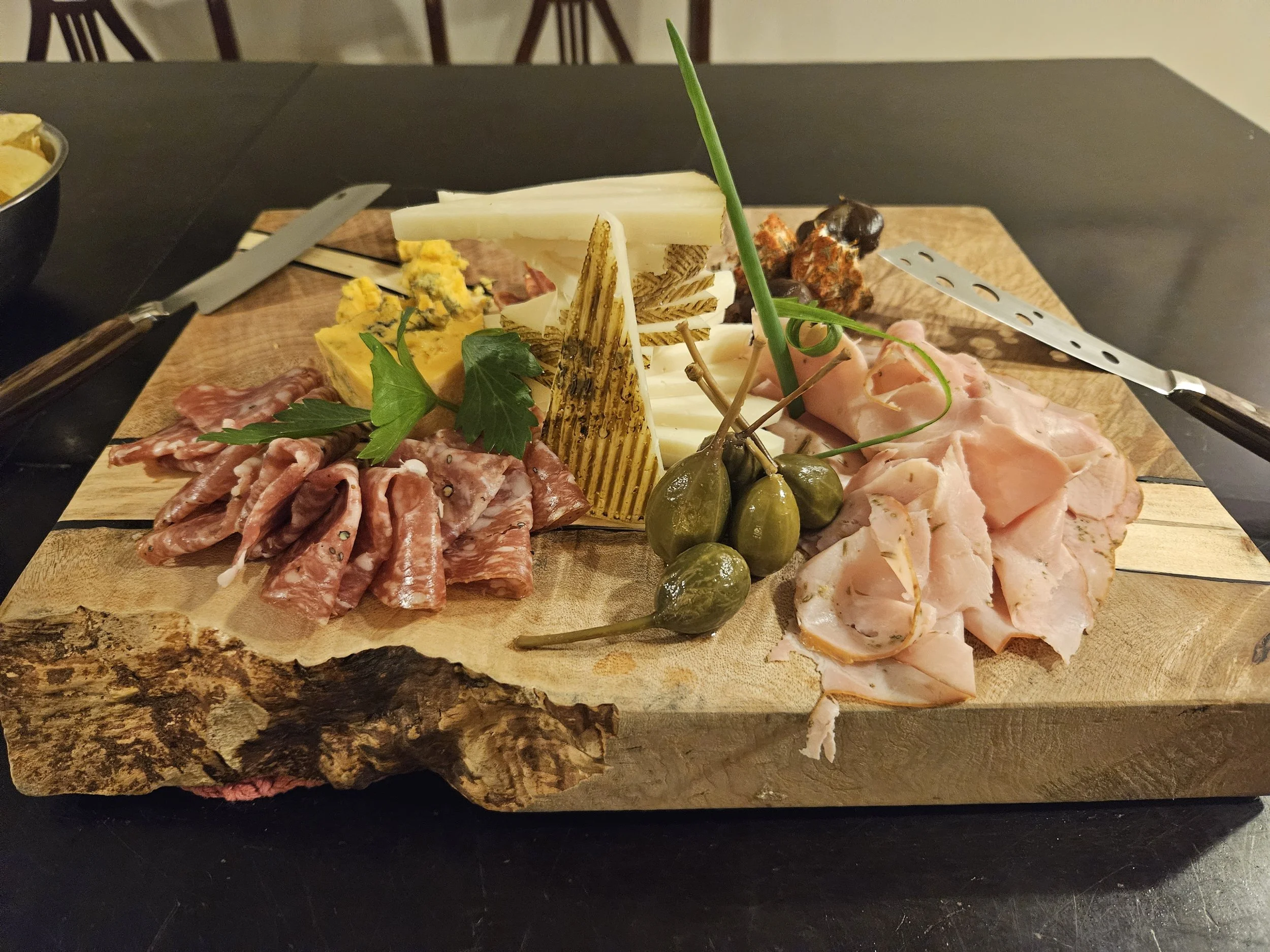 A wooden board with an assortment of cured meats, cheeses, pickles, and garnishes, served on a rustic chopping block. The meats include slices of prosciutto and salami, with cheese slices, green caperberries, and greenery for decoration.