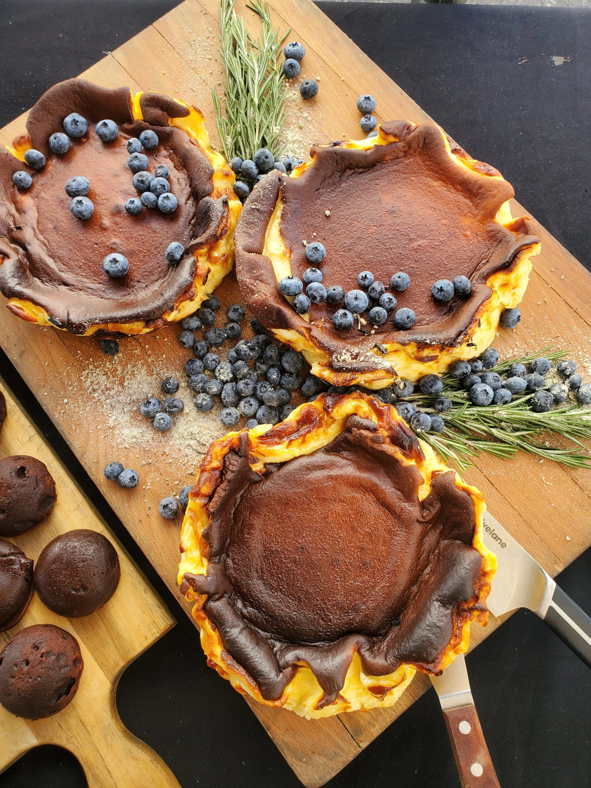 A yellow and brown baked cheesecake with a browned top, decorated with blueberries and sprigs of rosemary on a wooden cutting board.