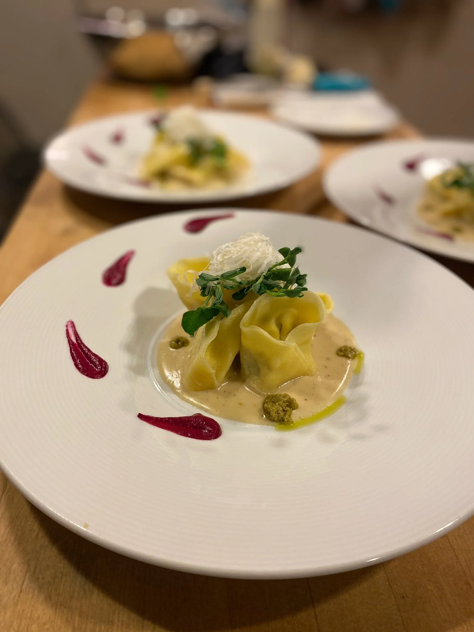Close-up of a plated dish with pasta, topped with a cheese, greens, and sauce on a white plate with red and purple decorative sauce highlights.