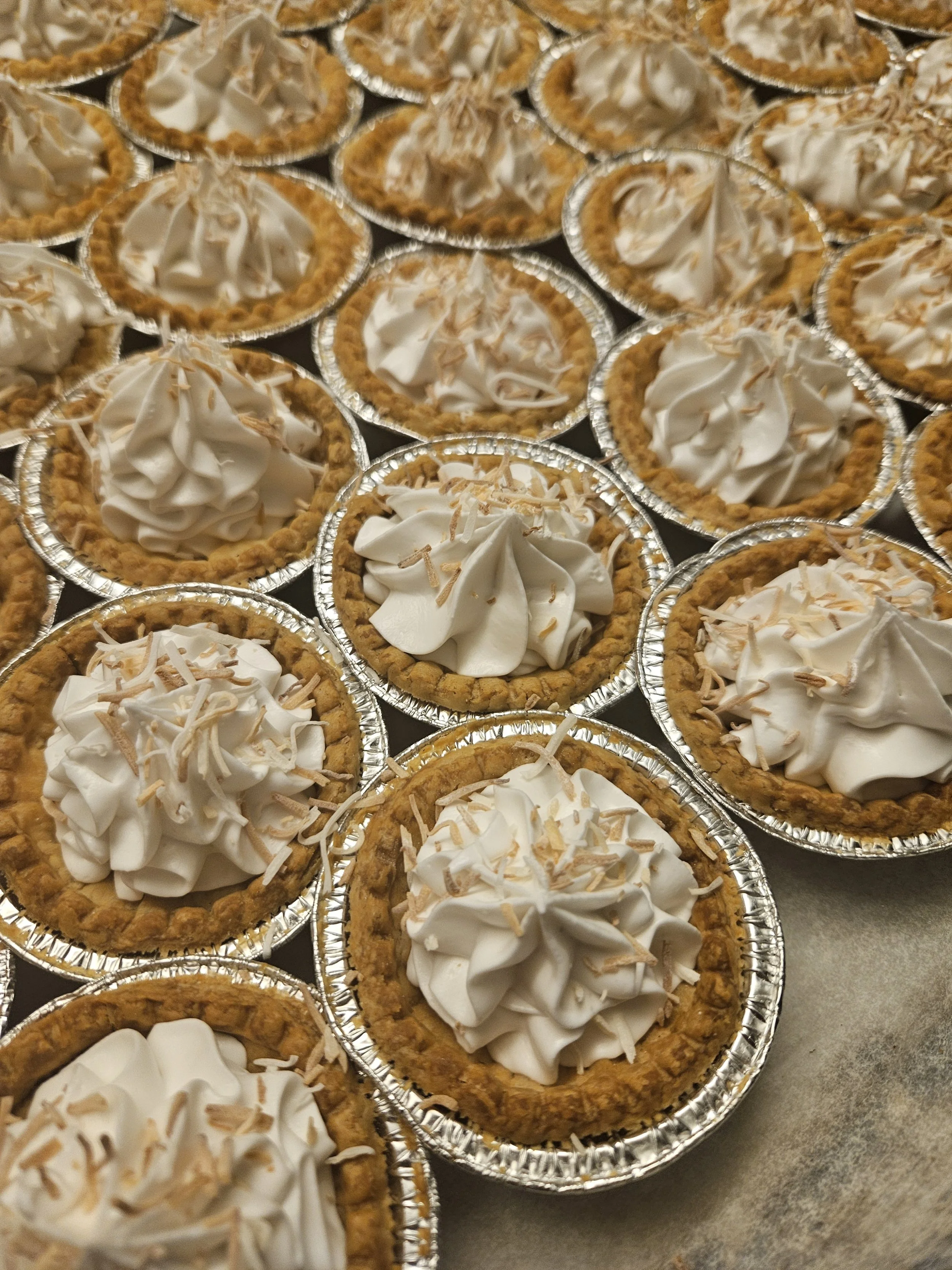 Multiple mini pies topped with whipped cream and toasted almond slices in aluminum pie tins on a countertop.