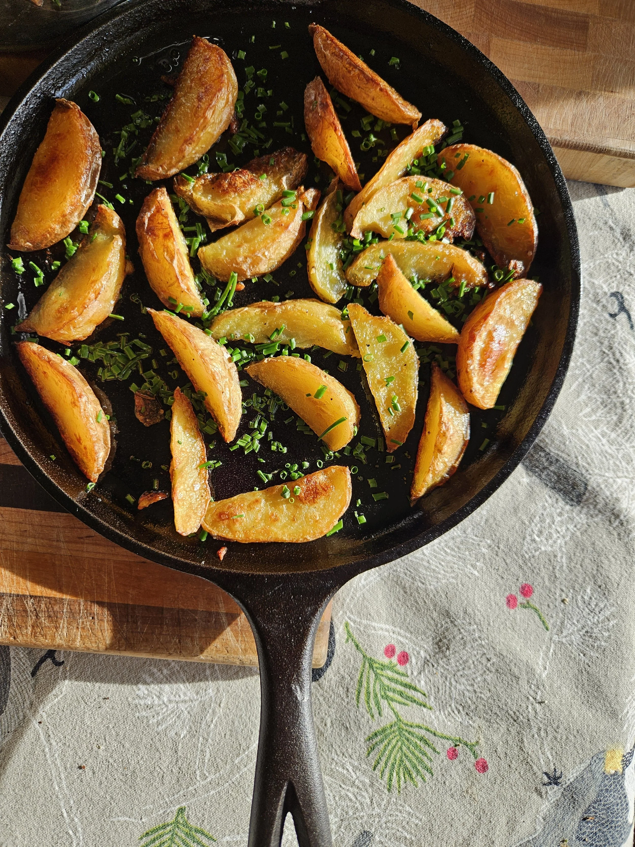 Cast iron skillet with roasted potato wedges garnished with chopped chives, placed on a wooden cutting board and a festive tablecloth with colorful berry and pine needle pattern.