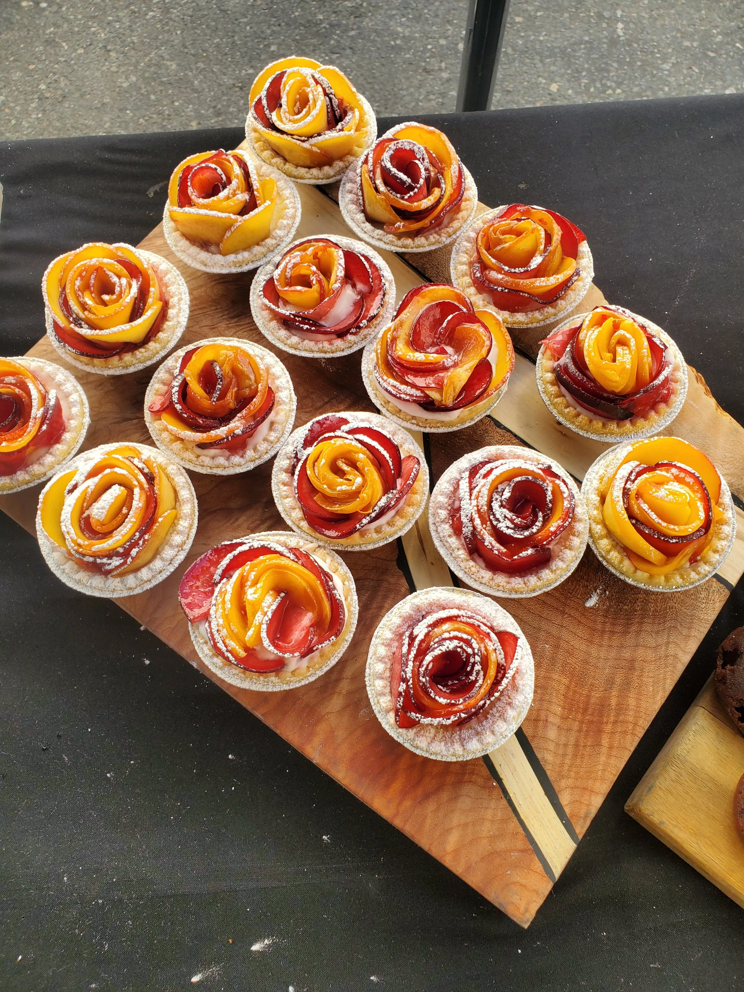 Small fruit tartlets shaped like roses with red, yellow, and orange fruit slices, dusted with powdered sugar, arranged on a wooden board.