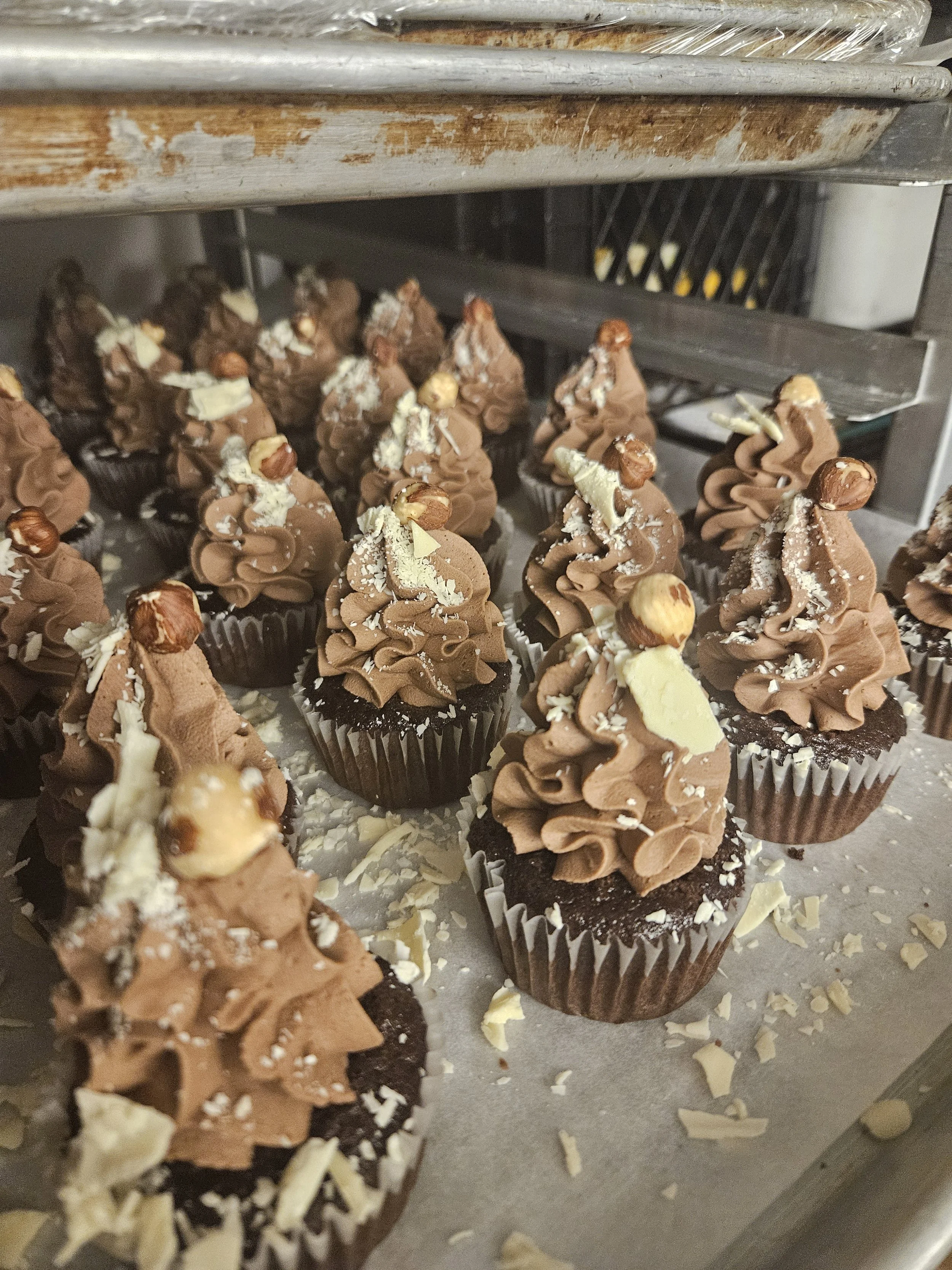 Chocolate cupcakes decorated with swirls of chocolate frosting, hazelnuts, and white chocolate shavings on a baking sheet.